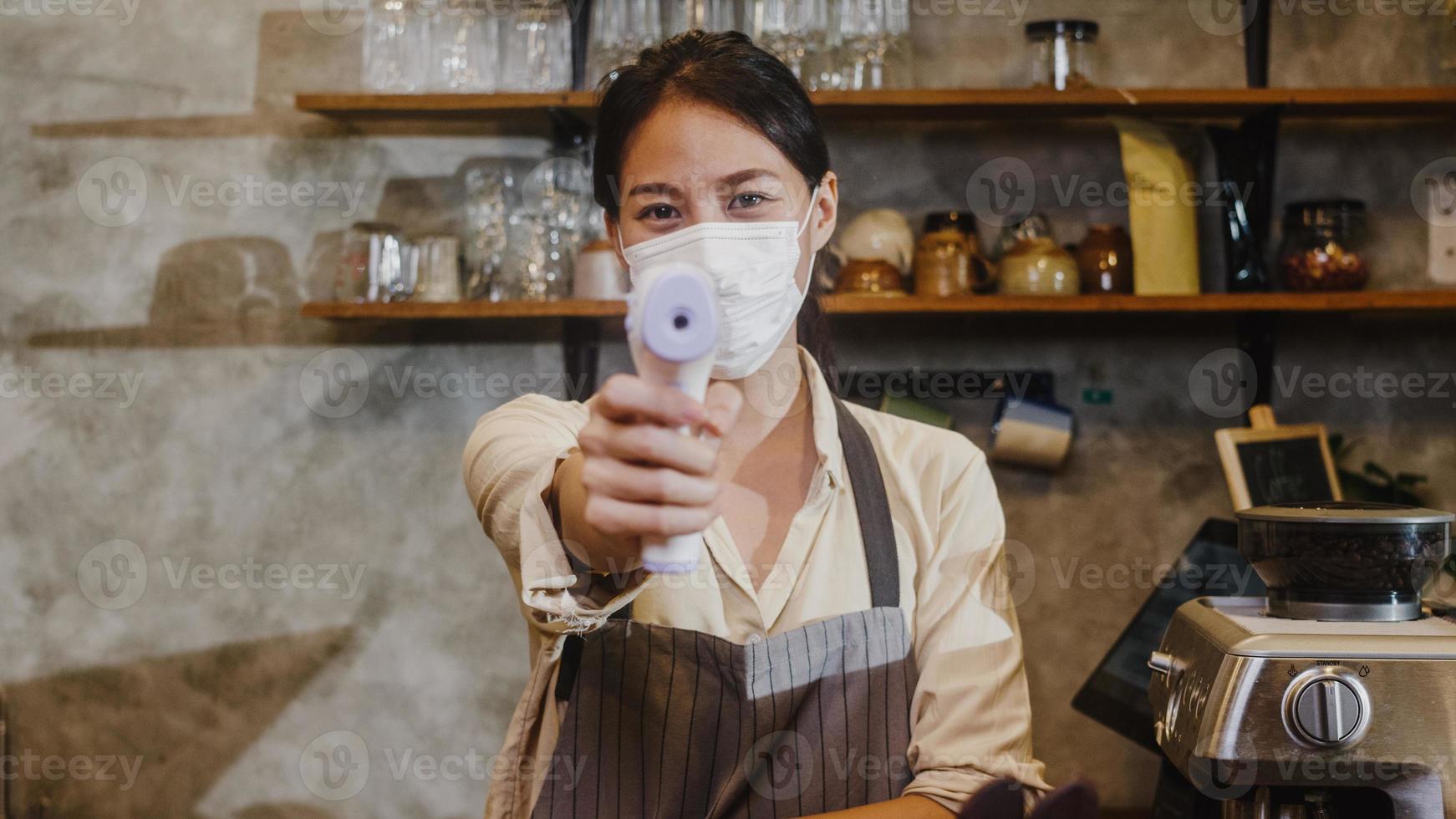 jonge Aziatische vrouwelijke restaurantmedewerkers die een beschermend gezichtsmasker dragen met behulp van een infraroodthermometercontrole of een temperatuurpistool op het voorhoofd van de klant voordat ze naar binnen gaan. levensstijl nieuw normaal na corona virus. foto