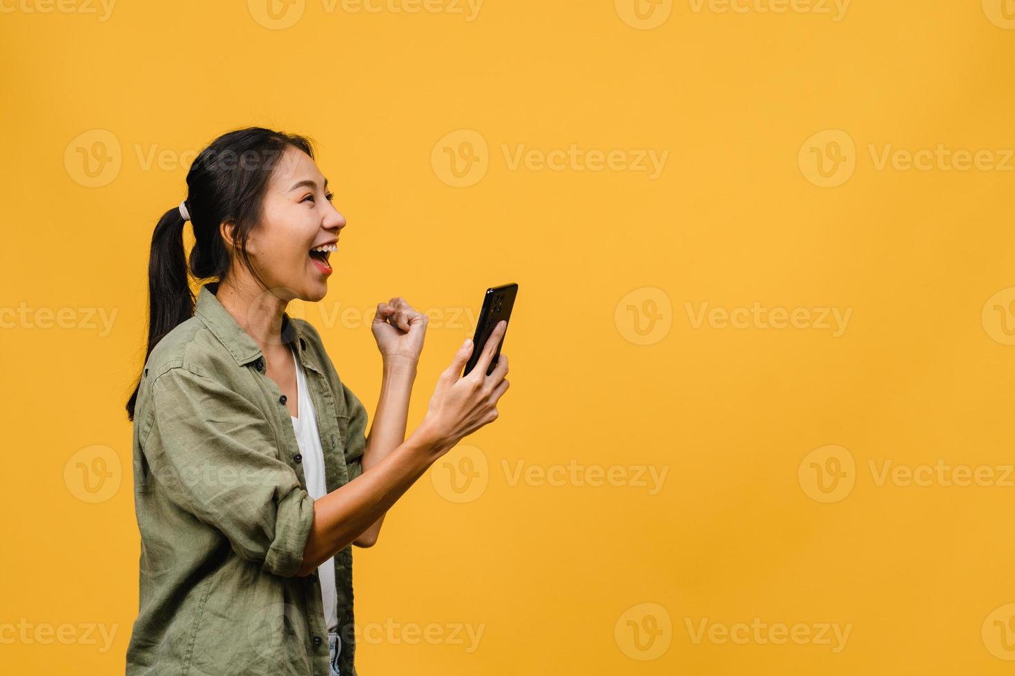 verraste jonge aziatische dame met behulp van mobiele telefoon met positieve uitdrukking, glimlacht breed, gekleed in casual kleding en staat geïsoleerd op gele achtergrond. gelukkige schattige blije vrouw verheugt zich over succes. foto