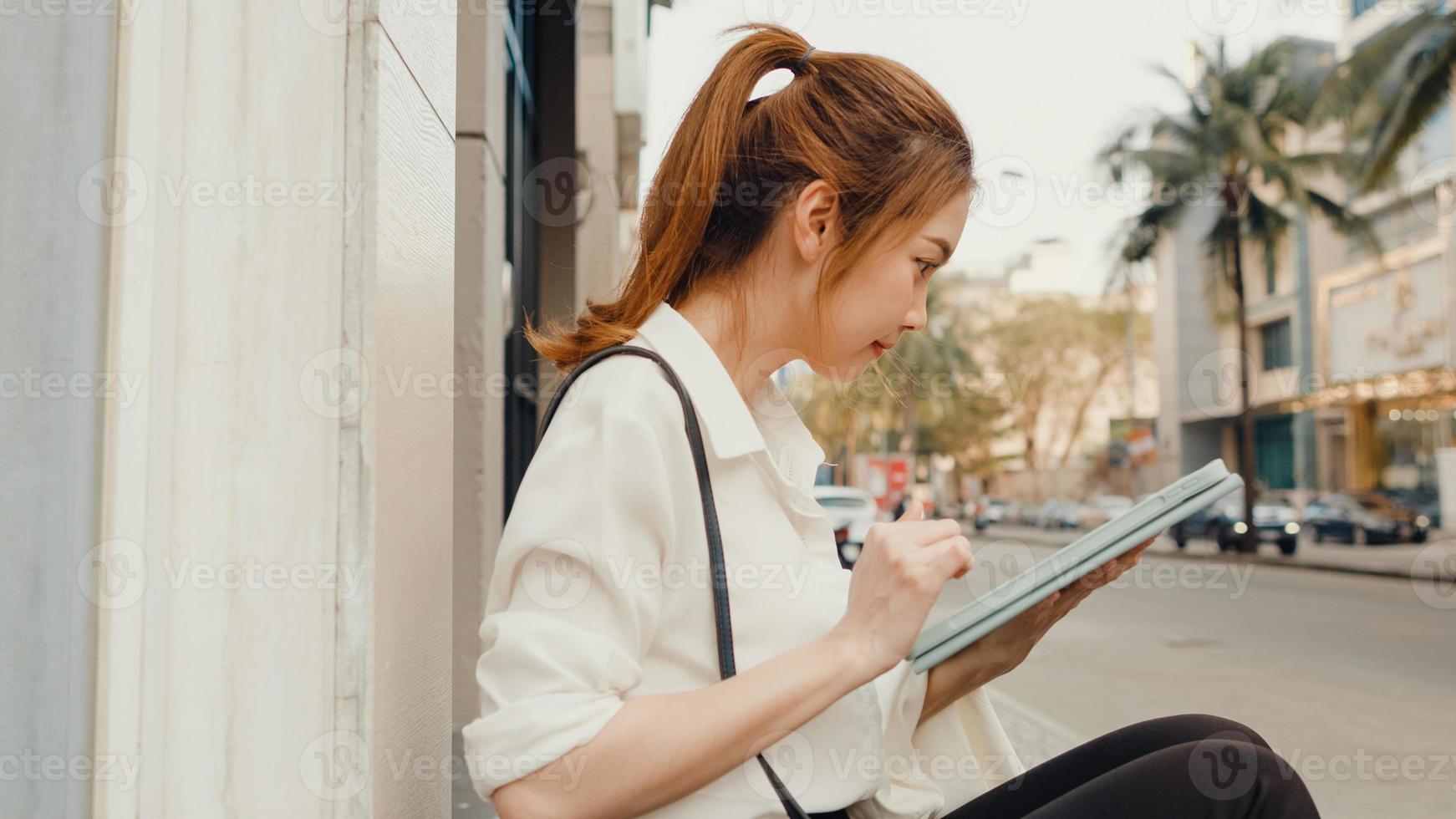 succesvolle jonge azië zakenvrouw in mode kantoorkleding met behulp van digitale tablet en tekstbericht typen terwijl ze 's ochtends alleen buiten in de stedelijke moderne stad zit. bedrijf onderweg concept. foto