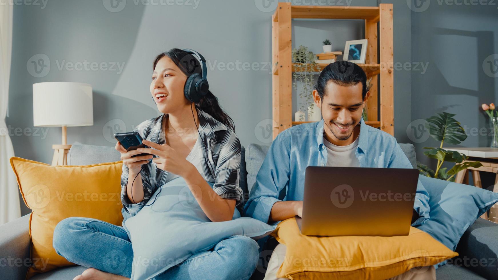 gelukkige jonge aziatische paar activiteit man gebruik laptop computer werk ontspannen genieten met vrouwen draag koptelefoon gebruik smartphone luister muziek op de bank in de woonkamer in huis. jong getrouwd werk vanuit huis concept. foto