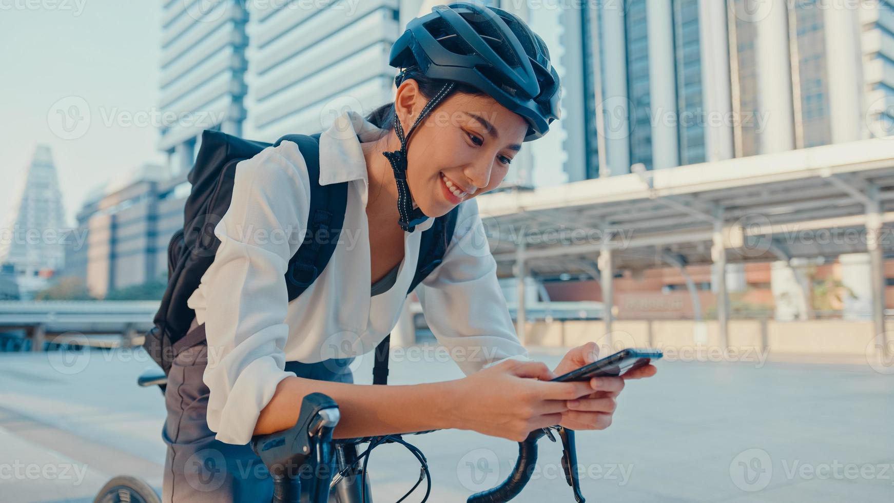 glimlach aziatische zakenvrouw met rugzak gebruik slimme telefoon kijk camera in stadsstand op straat met fiets ga aan het werk op kantoor. sport meisje telefoon gebruiken voor werk. pendelen naar het werk, zakelijke forens in de stad. foto