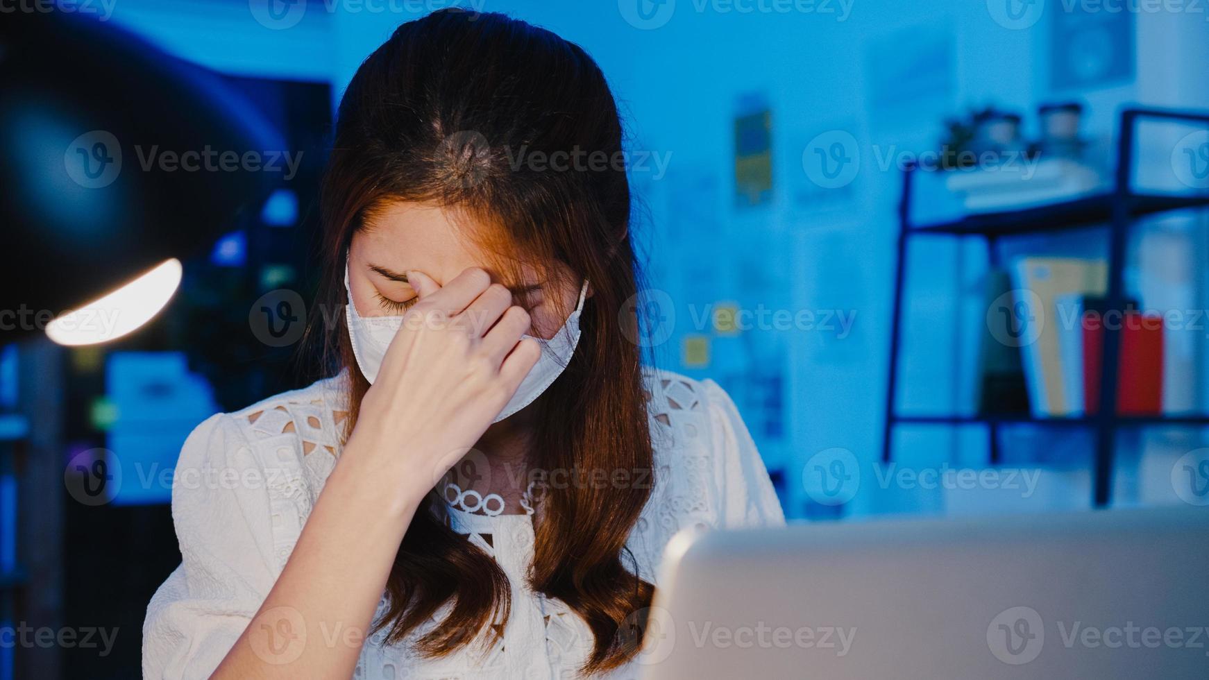 freelance azische vrouwen dragen gezichtsmasker met behulp van laptop hard werken op een nieuw normaal thuiskantoor. 's nachts werken vanuit huis overbelasting, zelfisolatie, sociale afstand, quarantaine voor preventie van het coronavirus. foto