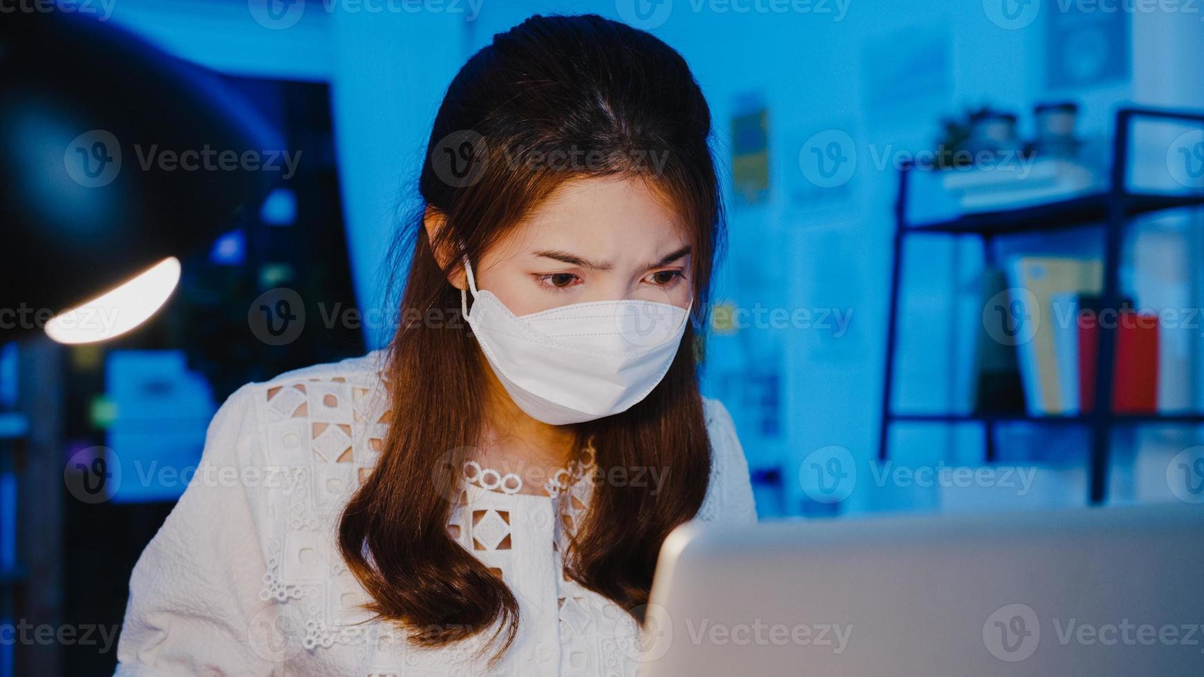 gelukkige zakenvrouw in azië die een medisch gezichtsmasker draagt voor sociale afstand in een nieuwe normale situatie voor viruspreventie tijdens het gebruik van een laptop op het werk in de kantoornacht. leven en werken na het coronavirus. foto