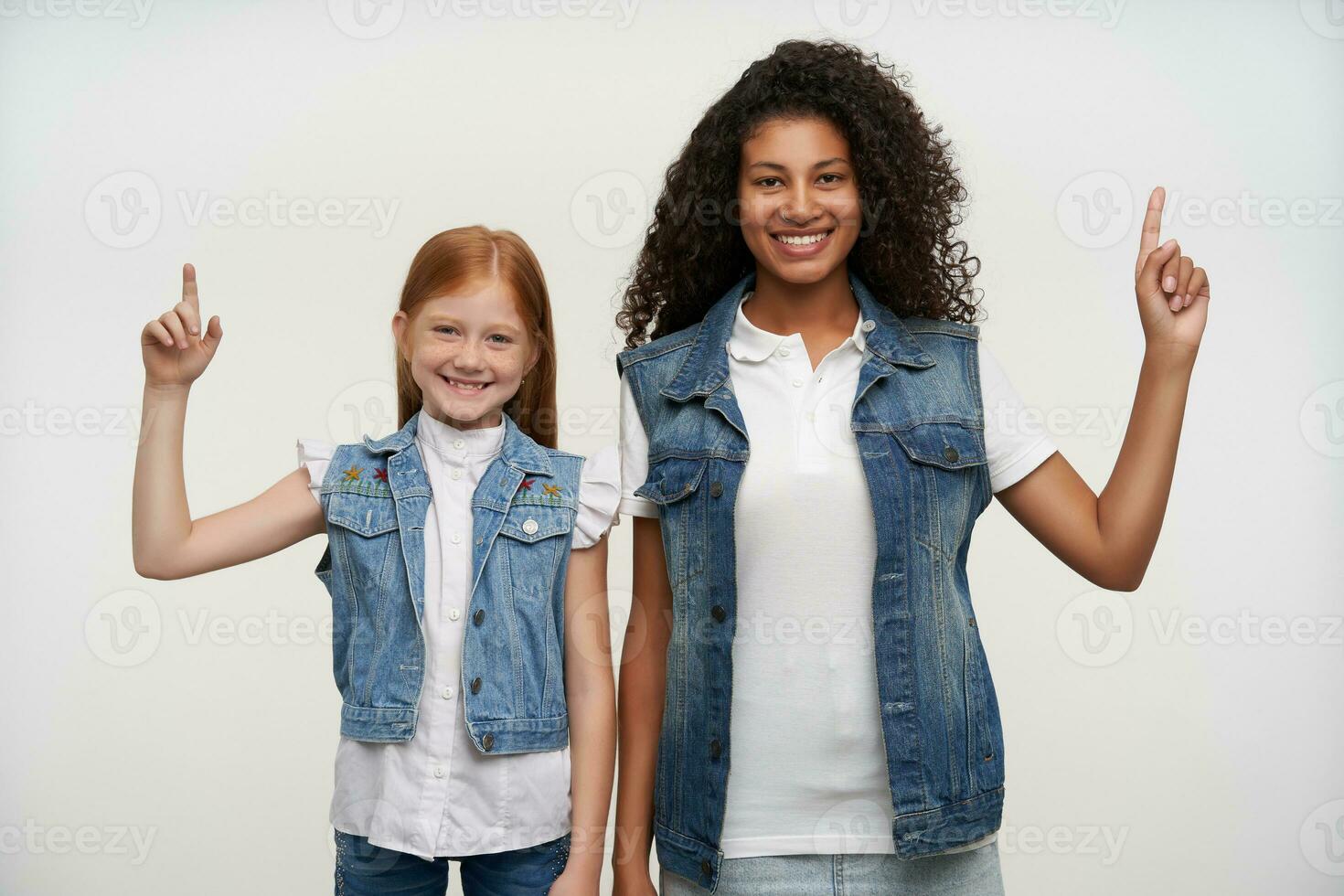 studio schot van vrolijk mooi jong meisjes met lang haar- verhogen omhoog hun wijsvingers en op zoek gelukkig naar camera met breed glimlacht, geïsoleerd over- wit achtergrond in gewoontjes kleren foto