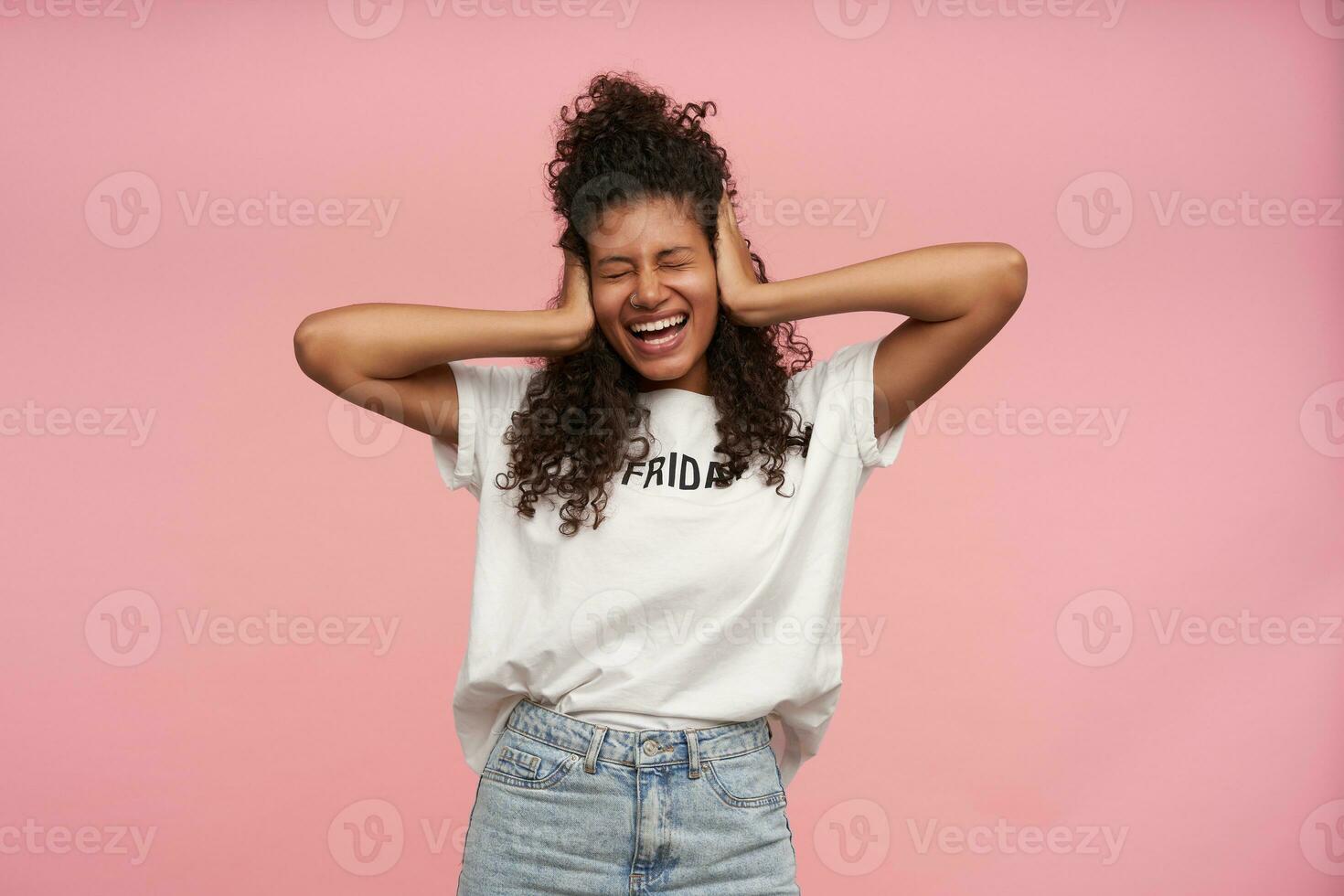 vrolijk aangenaam op zoek gekruld brunette vrouw met donker huid aan het bedekken haar oren met palmen en glimlachen blij met Gesloten ogen, staand over- roze achtergrond in wit t-shirt en blauw jeans foto