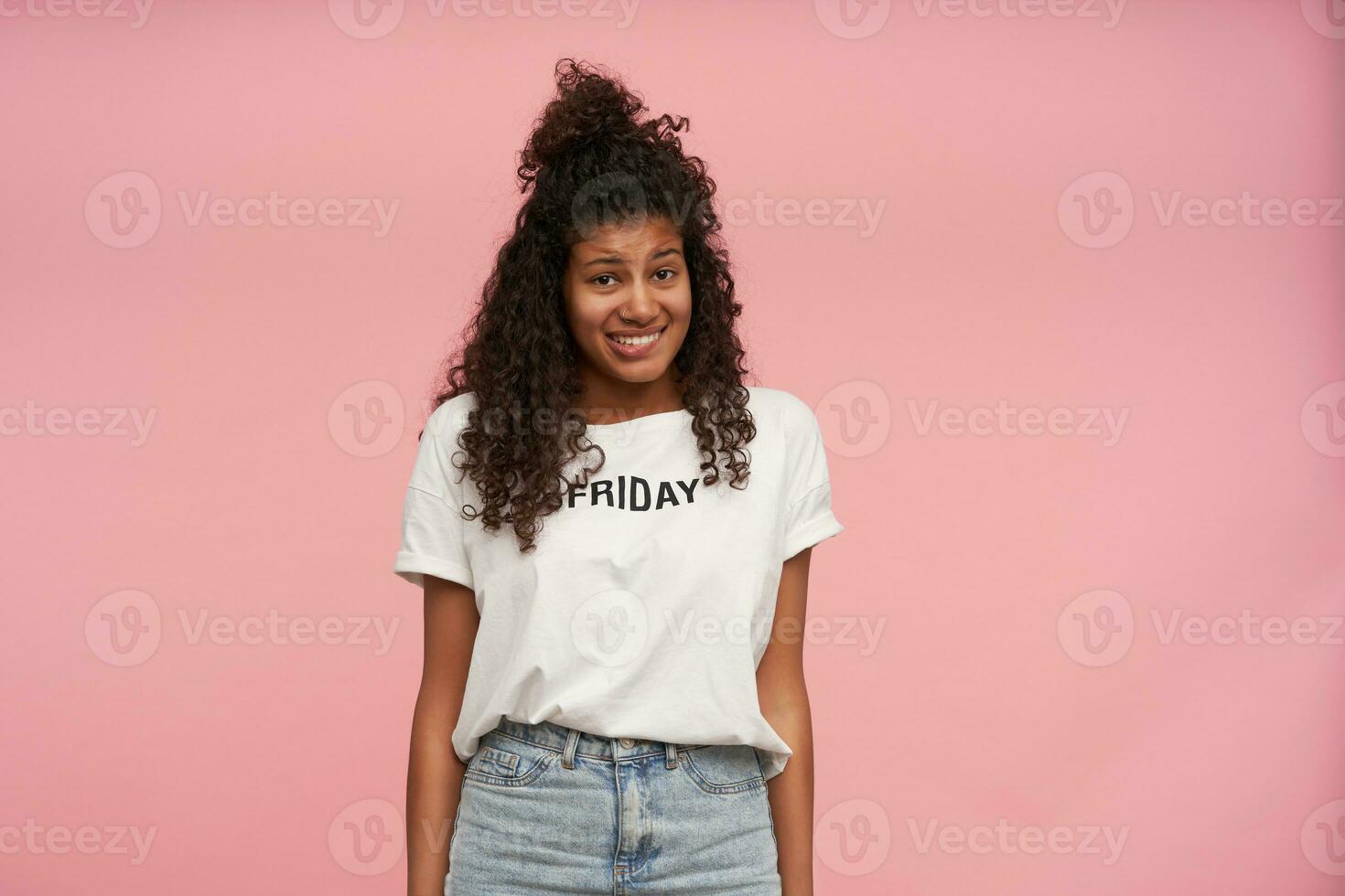 verward donker huid gekruld brunette vrouw op zoek Bij camera met oops gezicht en bijten onderlip, houden handen langs lichaam terwijl poseren over- roze achtergrond, gekleed in gewoontjes kleren foto