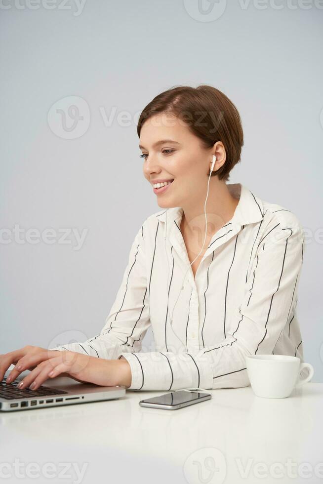 positief jong lief kort haren brunette dame luisteren naar muziek- in haar hoofdtelefoons terwijl typen tekst met toetsenbord Aan laptop, zittend over- wit achtergrond in gestreept overhemd foto