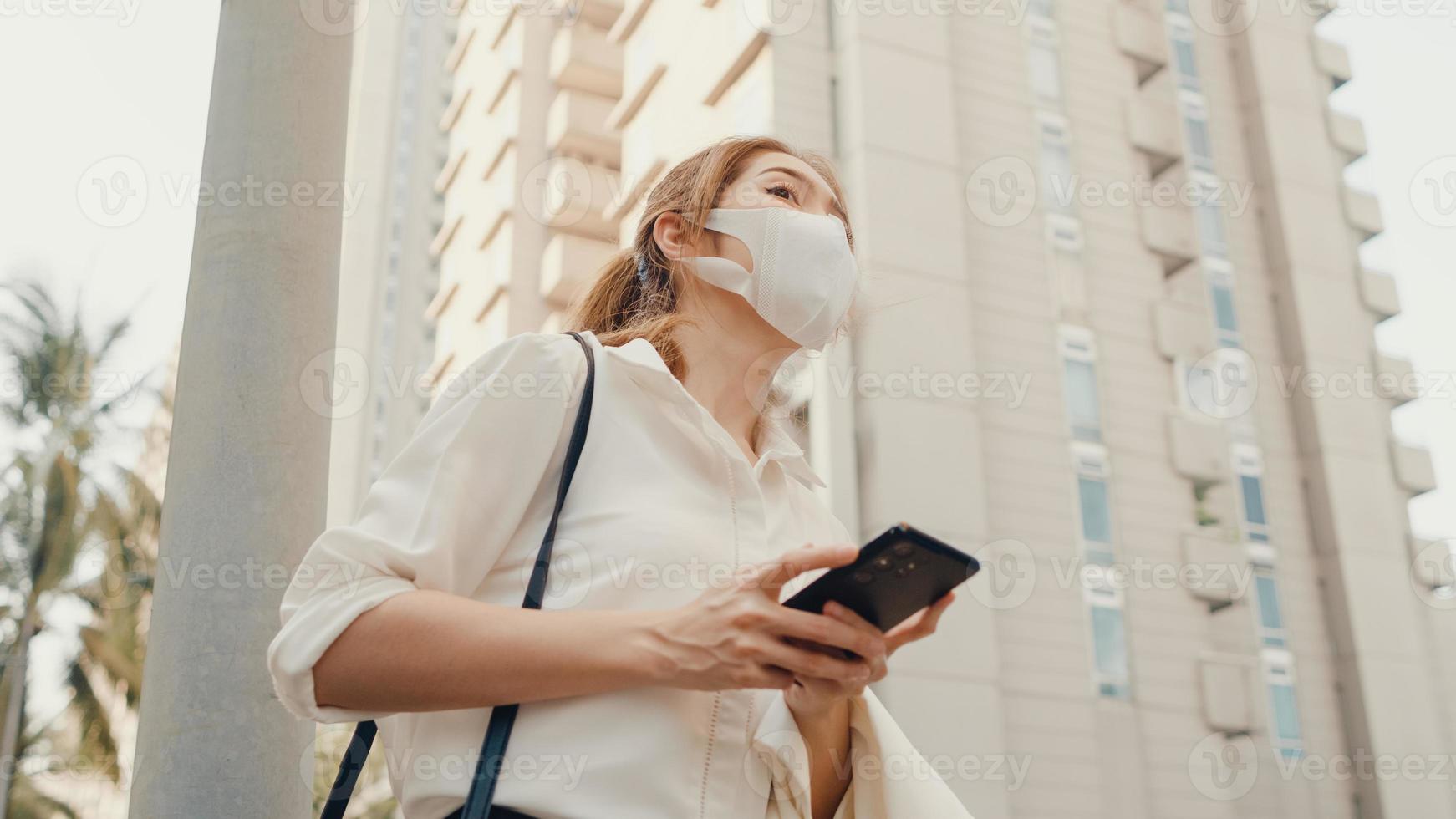 jonge Aziatische zakenvrouw in mode-kantoorkleding met een medisch gezichtsmasker dat op de weg een taxi neemt en een smartphone gebruikt terwijl ze buiten in de stedelijke moderne stad staat. bedrijf onderweg concept. foto