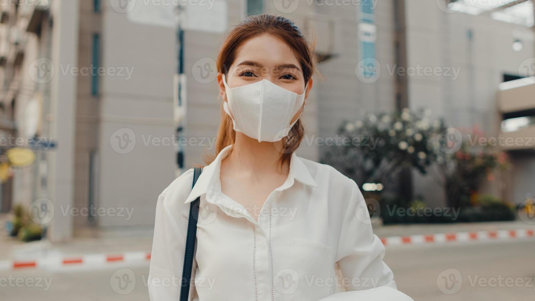 succesvolle jonge Aziatische zakenvrouw in mode-kantoorkleding draagt een medisch gezichtsmasker dat lacht en naar de camera kijkt terwijl ze gelukkig alleen staat buiten in de stedelijke moderne stad. bedrijf onderweg concept. foto