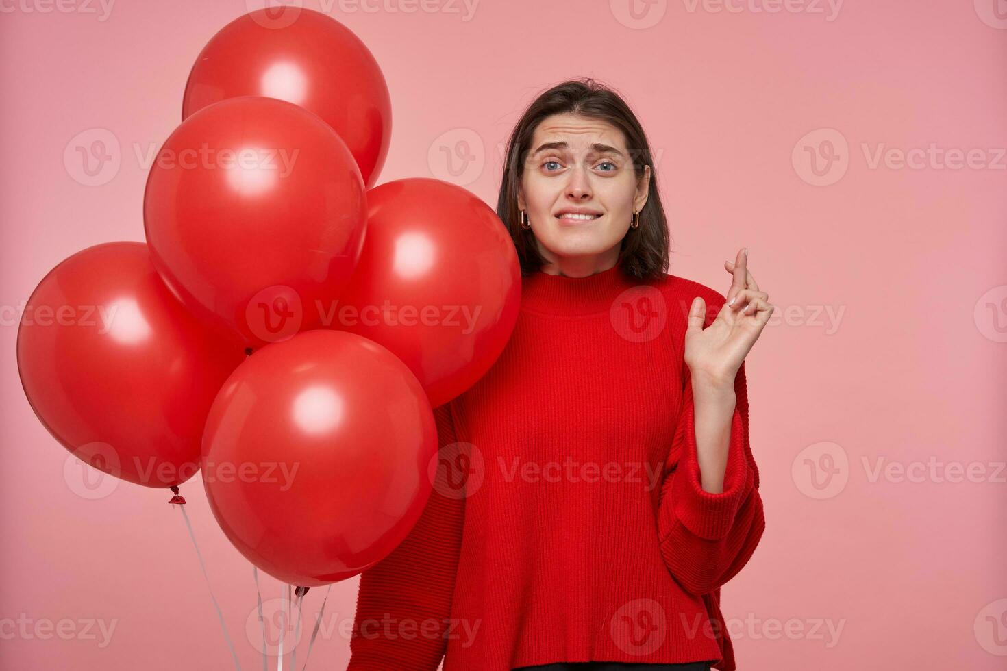 binnen- portret van jong brunette vrouw, draagt helder rood trui, gekruiste haar vinger, bijten haar lip terwijl bidden voor mooi zo geschenk. geïsoleerd over- roze achtergrond foto