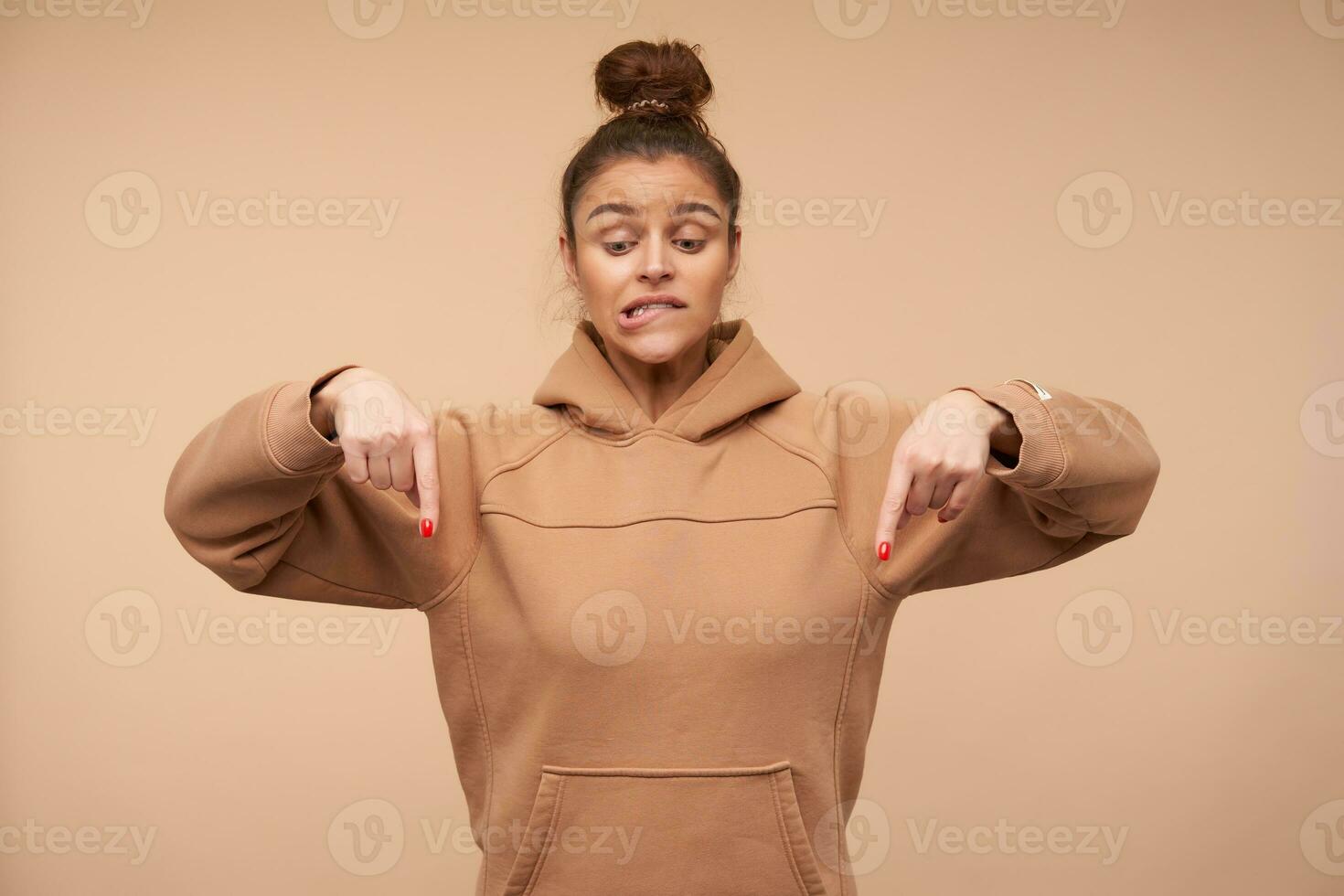 verbijsterd jong lief brunette vrouw met natuurlijk bedenken bijten zorgwekkend haar onderlip terwijl richten naar beneden met wijsvingers, staand over- beige achtergrond foto