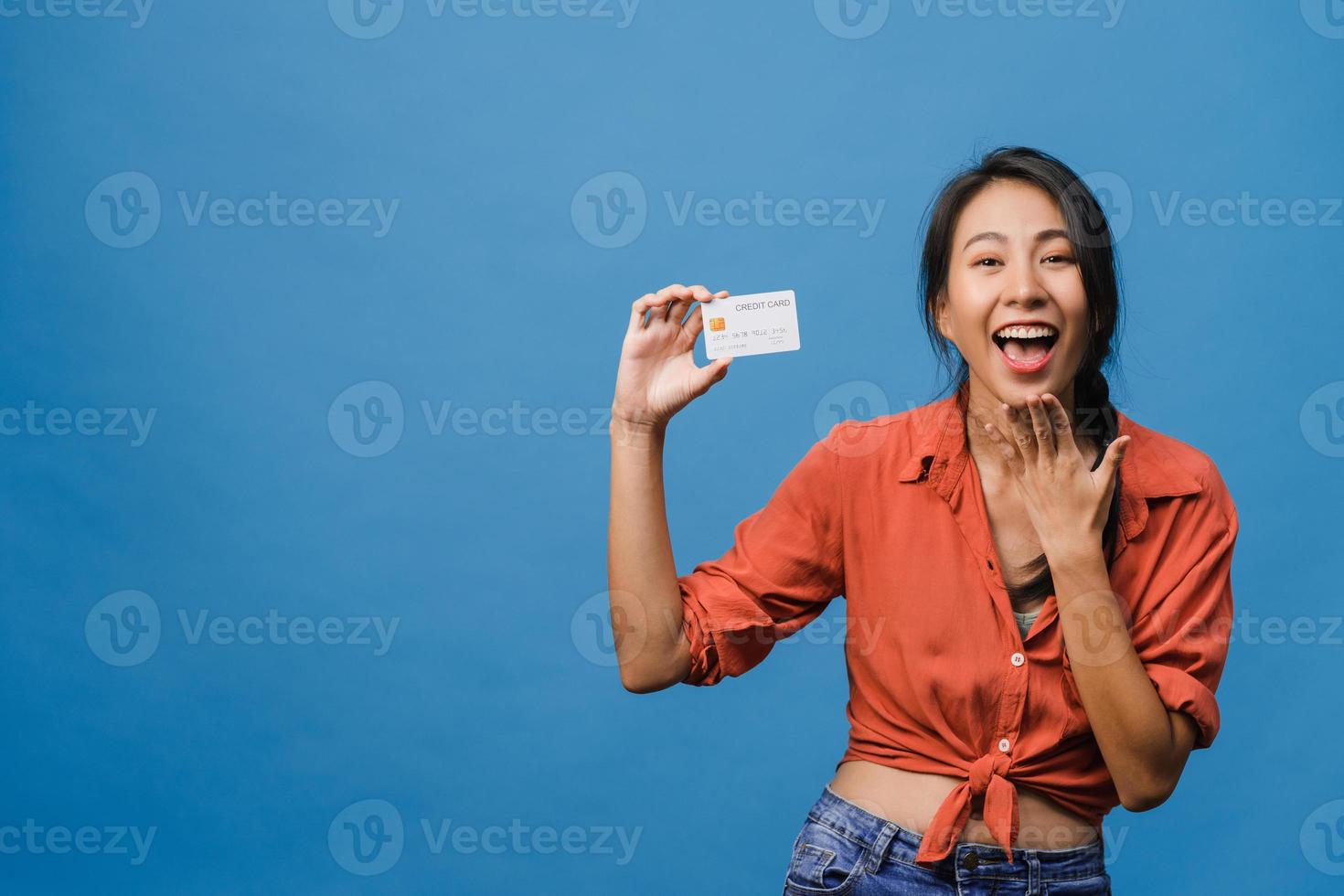 jonge aziatische dame toont creditcard met positieve uitdrukking, glimlacht breed, gekleed in casual kleding die geluk voelt en staat geïsoleerd op blauwe achtergrond. gezichtsuitdrukking concept. foto