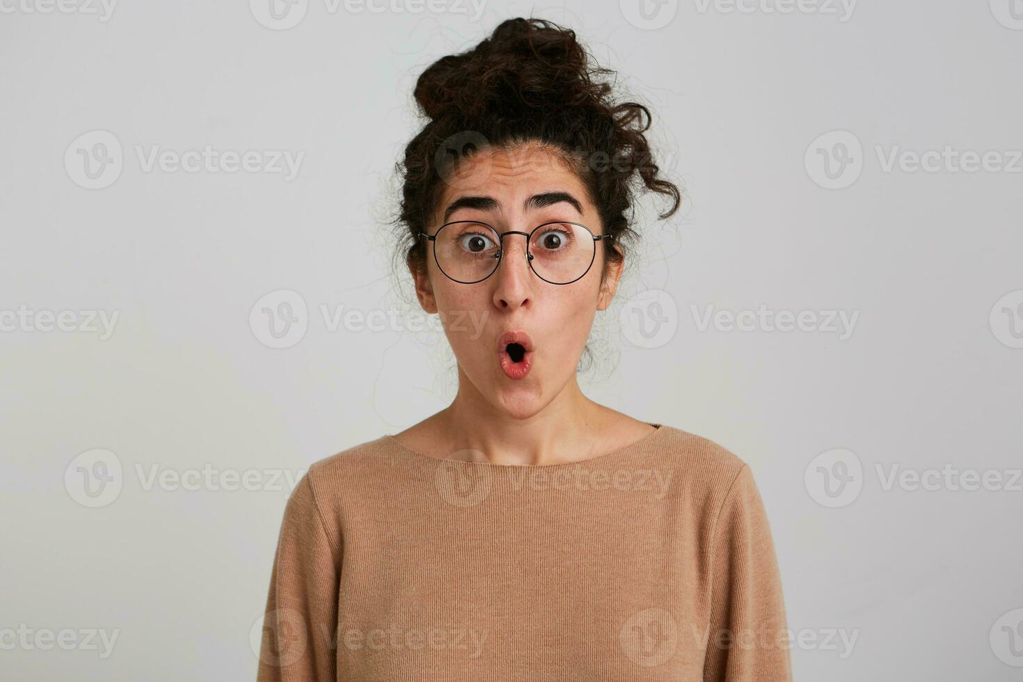 verrast op zoek vrouw, mooi meisje met donker gekruld haar- broodje. vervelend beige jumper en bril. emotie concept. geschokt over wat ze ziet. aan het kijken Bij de camera geïsoleerd over- wit achtergrond foto