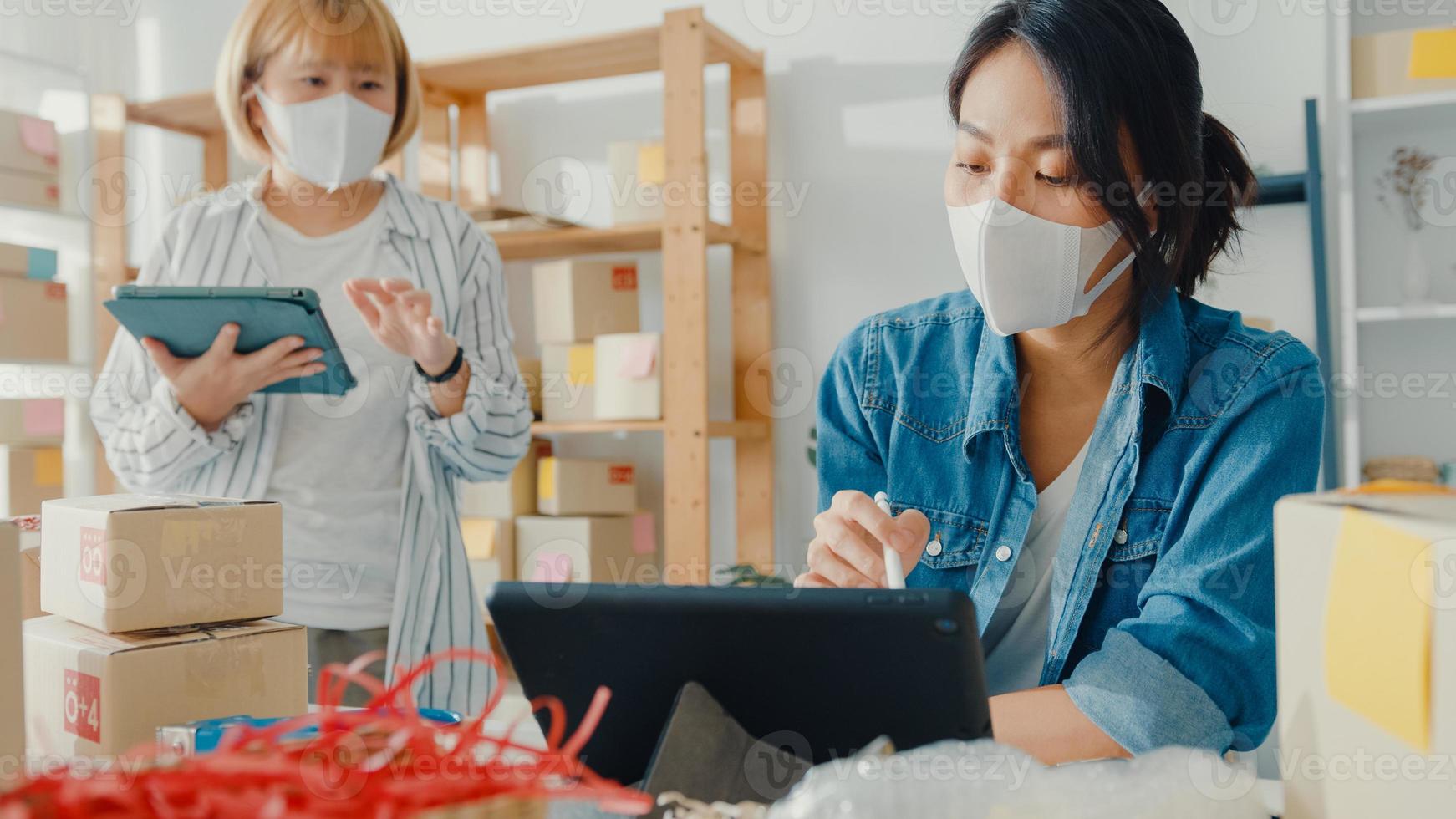 jonge Aziatische zakenvrouwen dragen een gezichtsmasker, controleren de aankooporder van het product op voorraad en bewaren ze op tabletcomputerwerk thuis. eigenaar van een klein bedrijf, online marktlevering, lifestyle freelance concept foto