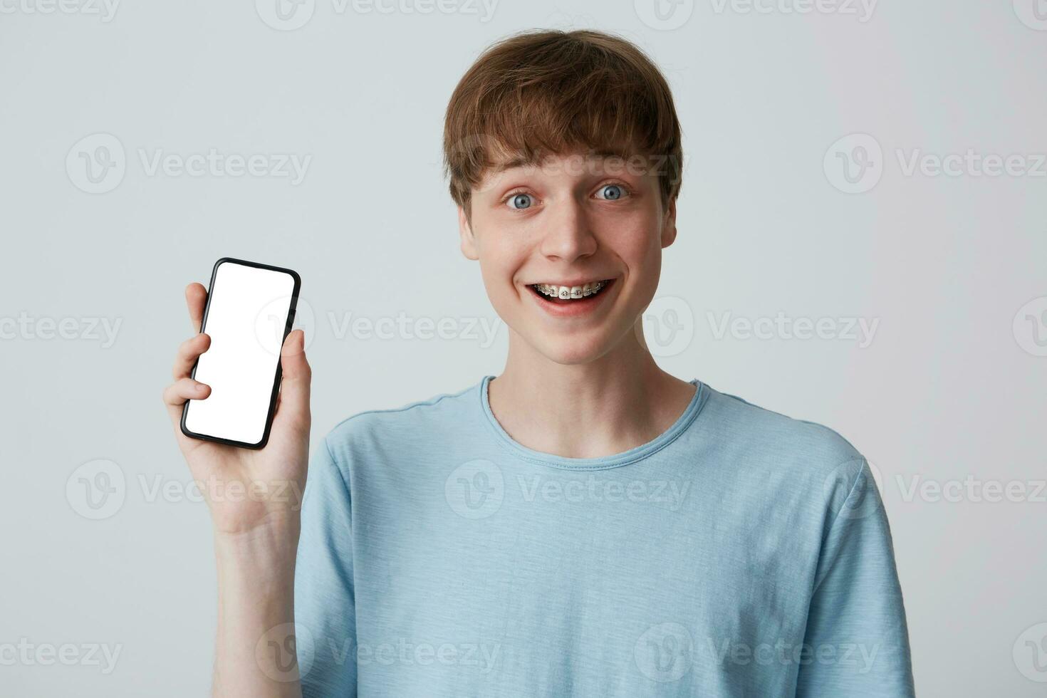 portret van vrolijk aantrekkelijk jong Mens leerling met een beugel Aan tanden draagt blauw t overhemd Holding blanco kopiëren ruimte scherm mobiel telefoon geïsoleerd over- wit achtergrond foto