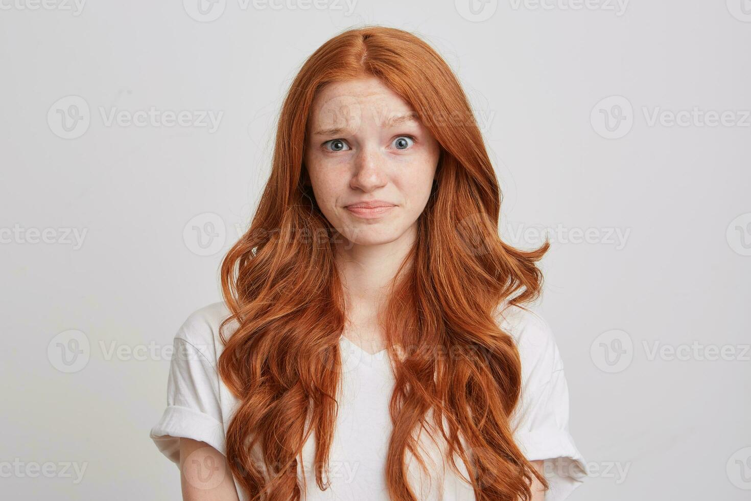 portret van verward verbaasd jong vrouw met lang golvend rood haar- en sproeten draagt t overhemd voelt in verlegenheid gebracht en looks direct in camera geïsoleerd over- wit achtergrond foto