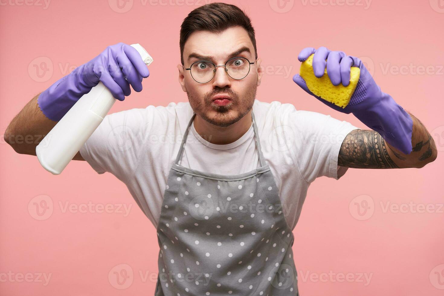 opgewonden jong mooi kort haren brunette Mens in bril verhogen verbaasd zijn wenkbrauwen en achtervolgen lippen terwijl Holding huishouden Chemicaliën in verheven handen, geïsoleerd over- roze achtergrond foto