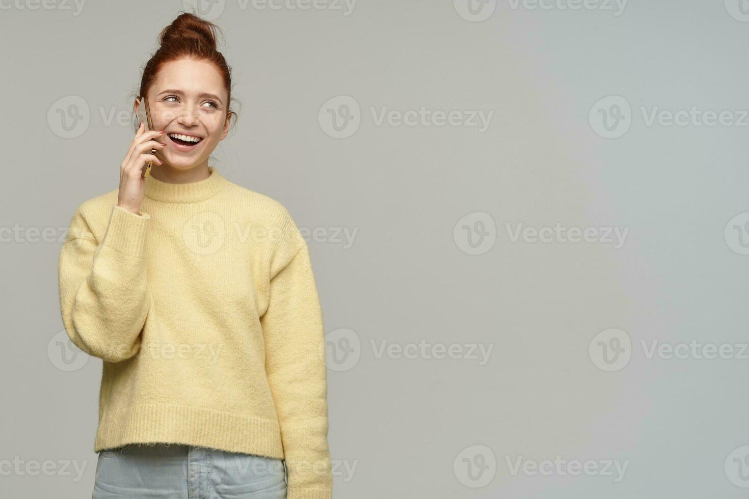 portret van aantrekkelijk, vrolijk meisje met gember haar- broodje. vervelend geel trui en pratend een telefoon. aan het kijken en lachend naar de Rechtsaf Bij kopiëren ruimte, geïsoleerd over- grijs achtergrond foto