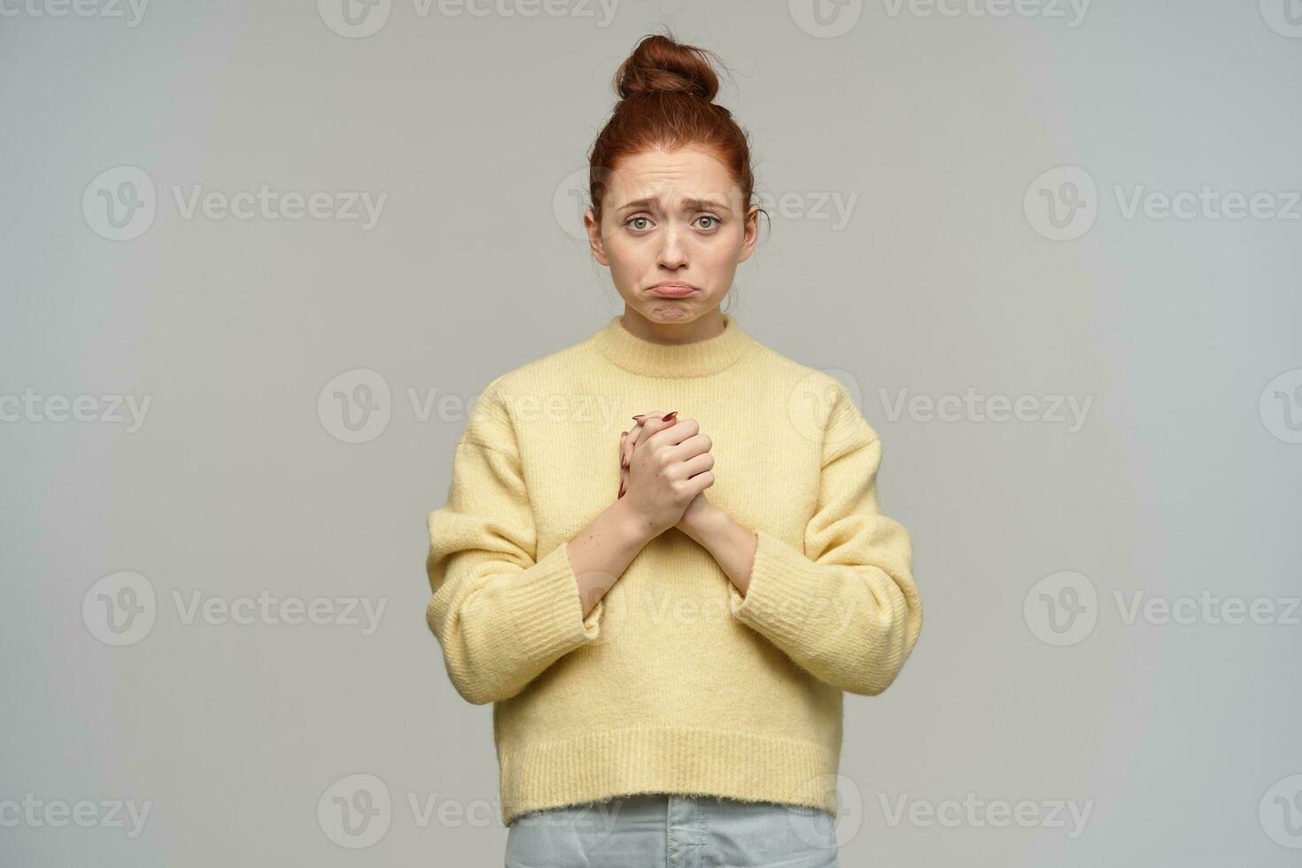 tiener- meisje, van streek op zoek vrouw met gember haar- verzameld in een broodje. vervelend pastel geel trui en jeans. houden armen samen, voelt sorry. aan het kijken Bij de camera geïsoleerd over- grijs achtergrond foto