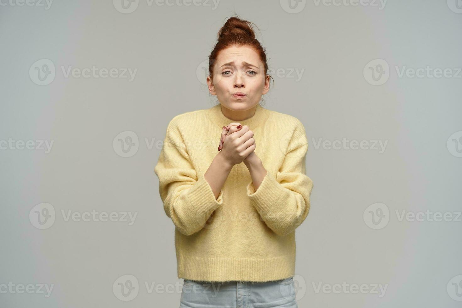 schattig op zoek vrouw, ten einde raad meisje met gember haar- verzameld in een broodje. vervelend pastel geel trui en jeans. houden haar armen samen, bedelen. aan het kijken Bij de camera geïsoleerd over- grijs achtergrond foto