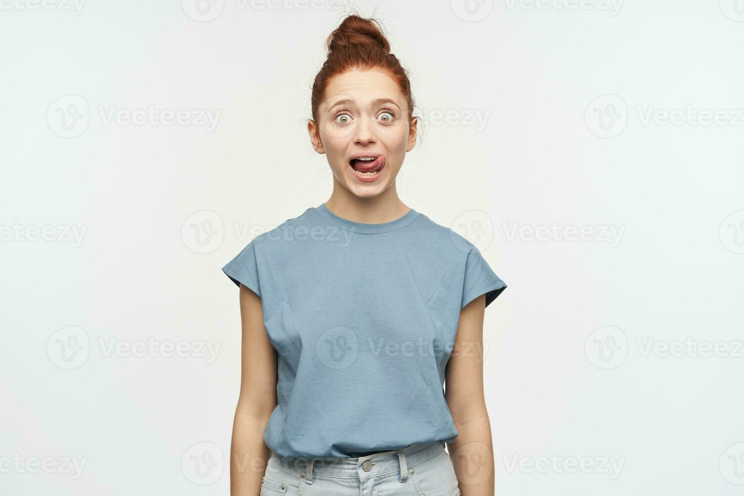 geschokt op zoek vrouw, mooi meisje met gember haar- verzameld in een broodje. vervelend blauw t-shirt en jeans. likken haar lip, shows tong. aan het kijken Bij de camera geïsoleerd over- wit achtergrond foto