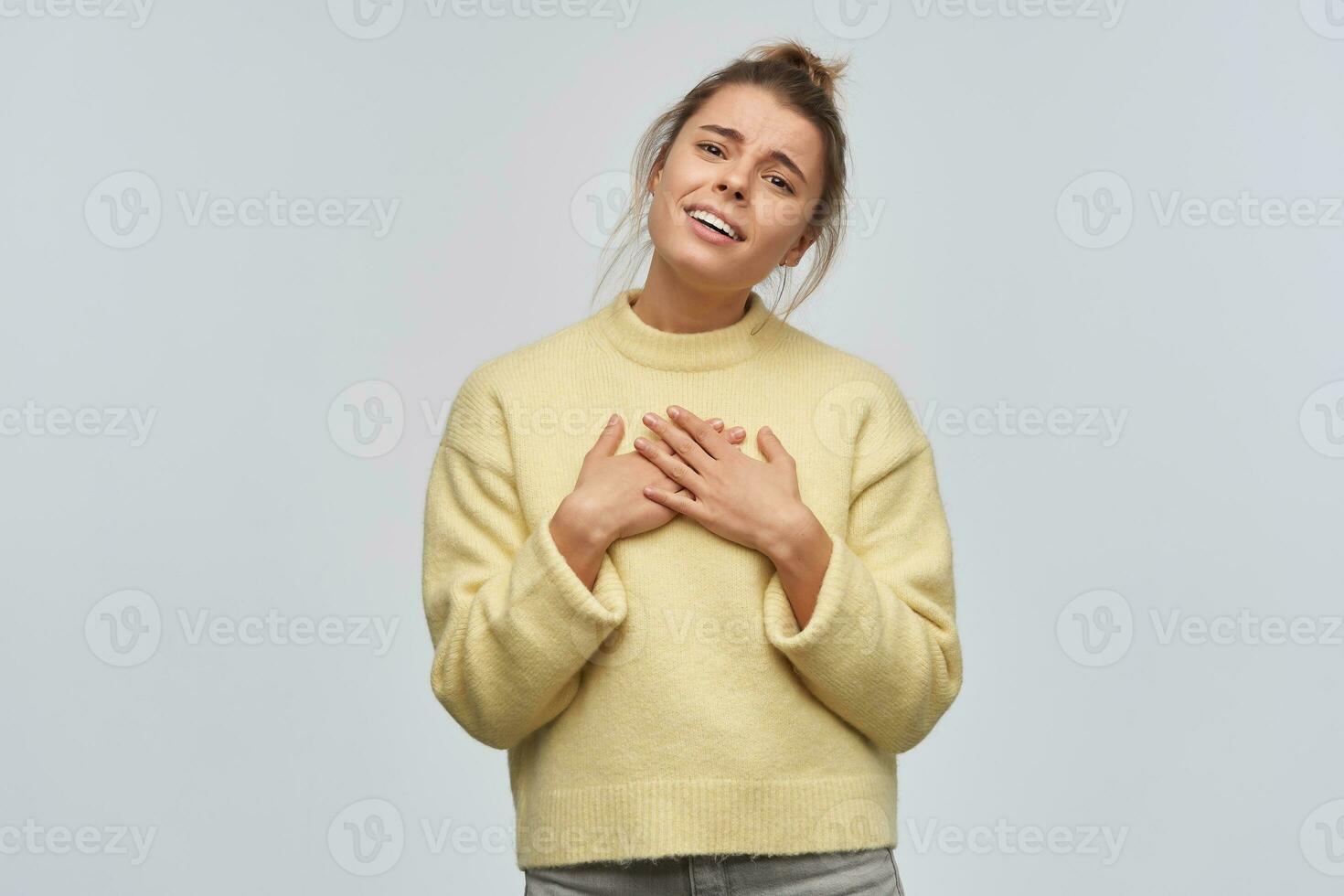 tiener- meisje, charmant vrouw met blond haar- verzameld in broodje. vervelend geel trui. kantelen haar hoofd en zet palmen Aan haar borst. horen compliment. aan het kijken Bij de camera, geïsoleerd over- wit achtergrond foto