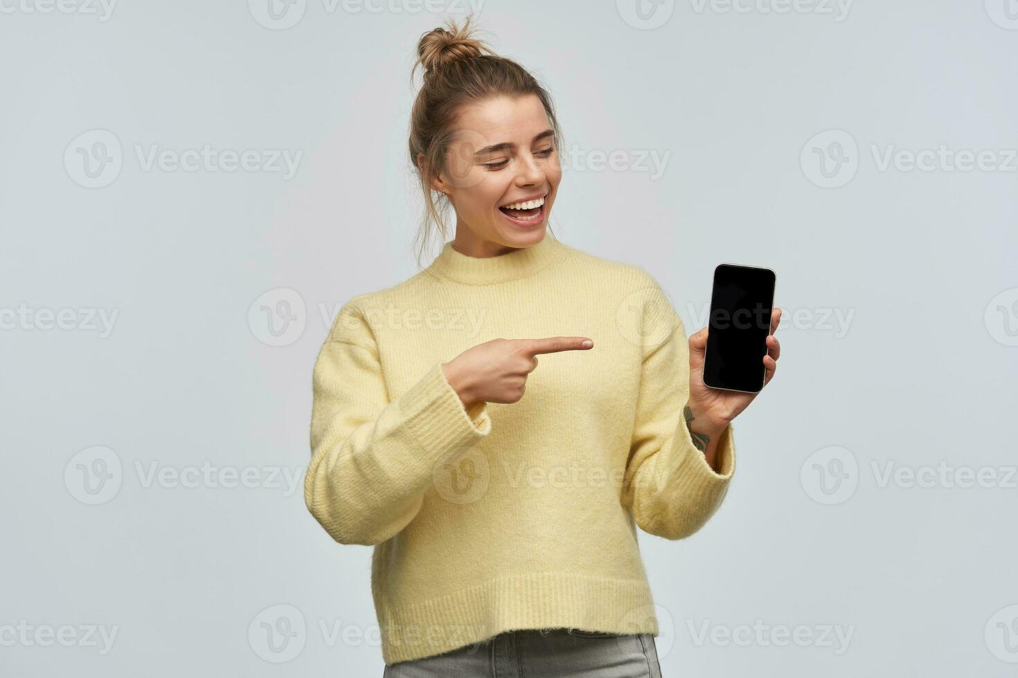 tiener- meisje, gelukkig op zoek vrouw met blond haar- verzameld in broodje. vervelend geel trui. aan het kijken en points naar de telefoon scherm, kopiëren ruimte. staan geïsoleerd over- wit achtergrond foto