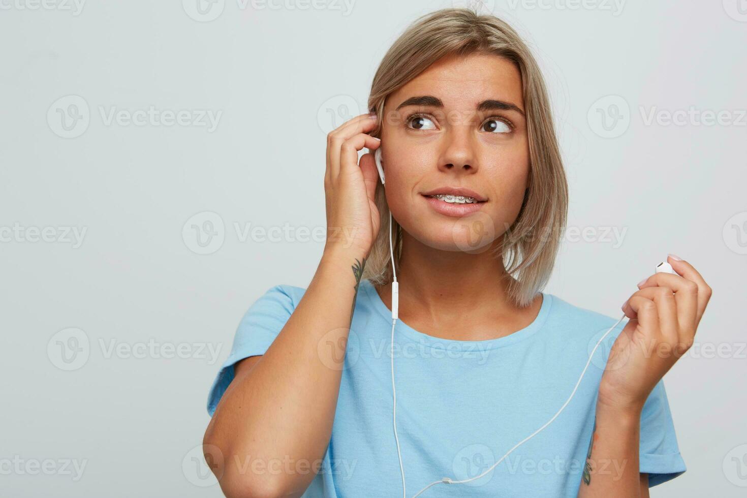 detailopname van gelukkig aantrekkelijk blond jong vrouw met een beugel Aan tanden en oortelefoons draagt blauw t overhemd looks ontspannen en luisteren naar muziek- geïsoleerd over- wit achtergrond foto