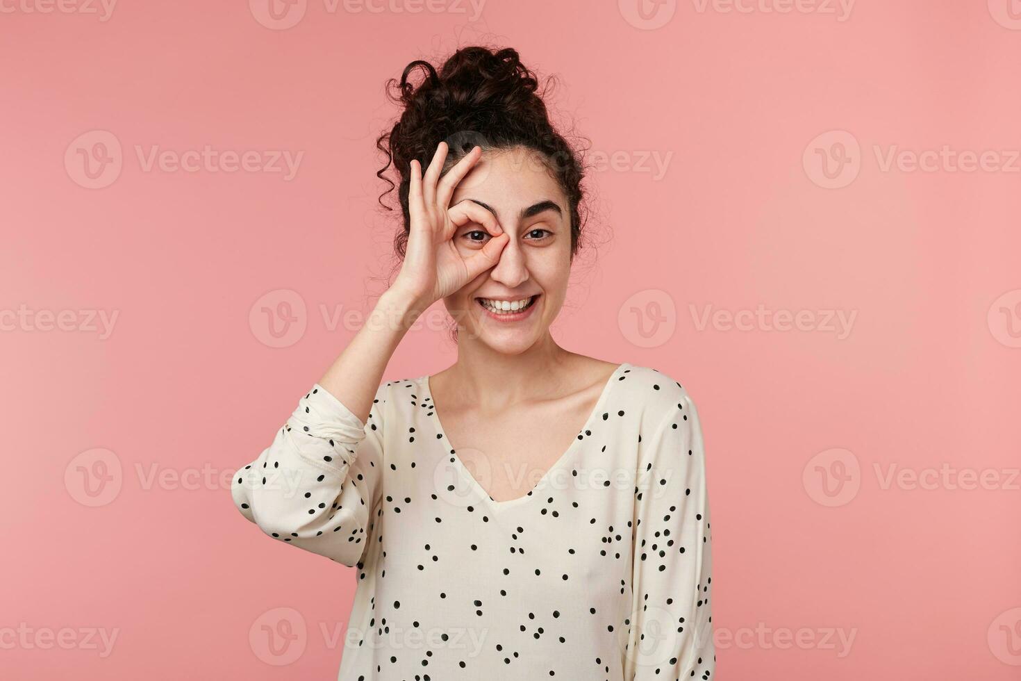 dichtbij omhoog portret van aantrekkelijk positief charmant brunette meisje met verzameld Golf haar- maken okey symbool dichtbij naar oog met een hand, in blouse met polka stippen, geïsoleerd Aan roze achtergrond foto