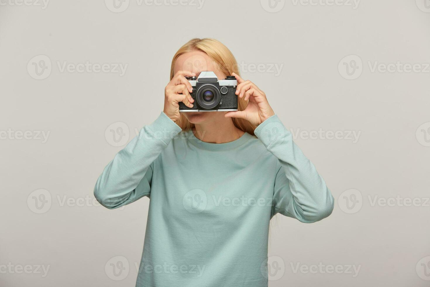 portret van een fotograaf aan het bedekken haar gezicht met de camera. fotograaf nemen foto, werken in studio, vervelend blauw gewoontjes trui, over- grijs achtergrond foto