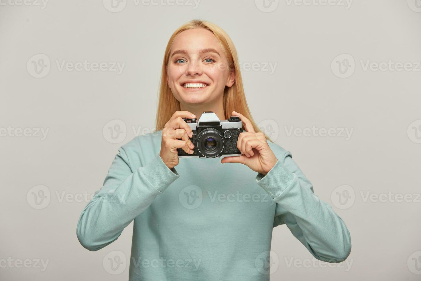 jong meisje fotograaf looks gelukkig lachend, Holding een retro wijnoogst foto camera in handen, in de werkwijze van schieten, werken in studio, vervelend blauw gewoontjes trui, over- grijs achtergrond