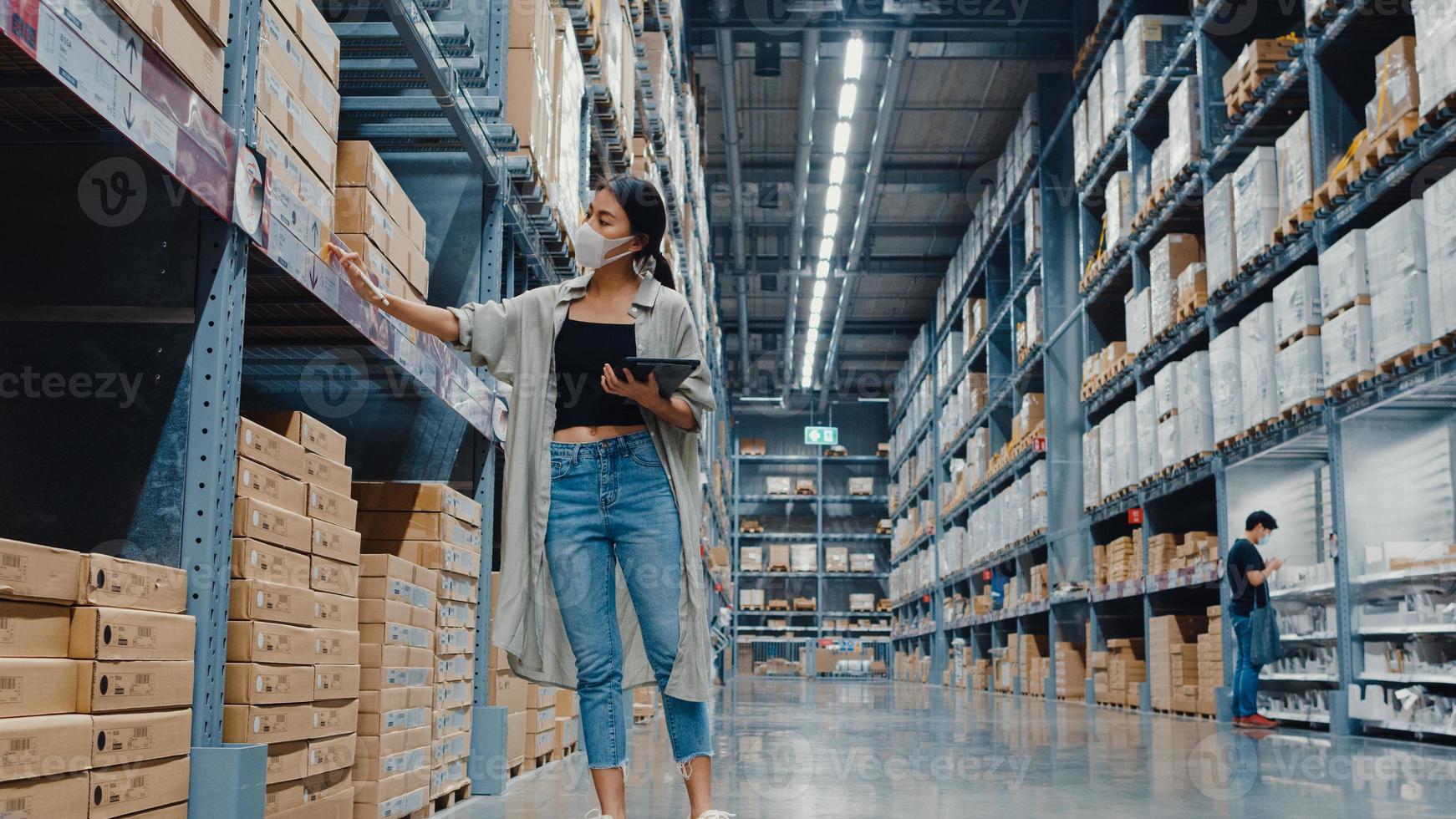 jonge azië zakenvrouw manager draagt gezichtsmasker op zoek naar goederen met behulp van digitale tablet controleren voorraadniveaus staan in retail winkelcentrum. distributie, logistiek, pakketten klaar voor verzending. foto