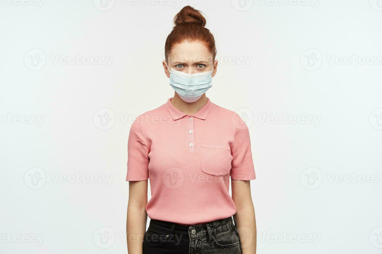 ongelukkig vrouw, fronsen roodharige meisje met haar- broodje. vervelend roze t-shirt en medisch, beschermend gezicht masker. staren negatief Bij jij. aan het kijken Bij de camera, geïsoleerd over- wit achtergrond. foto