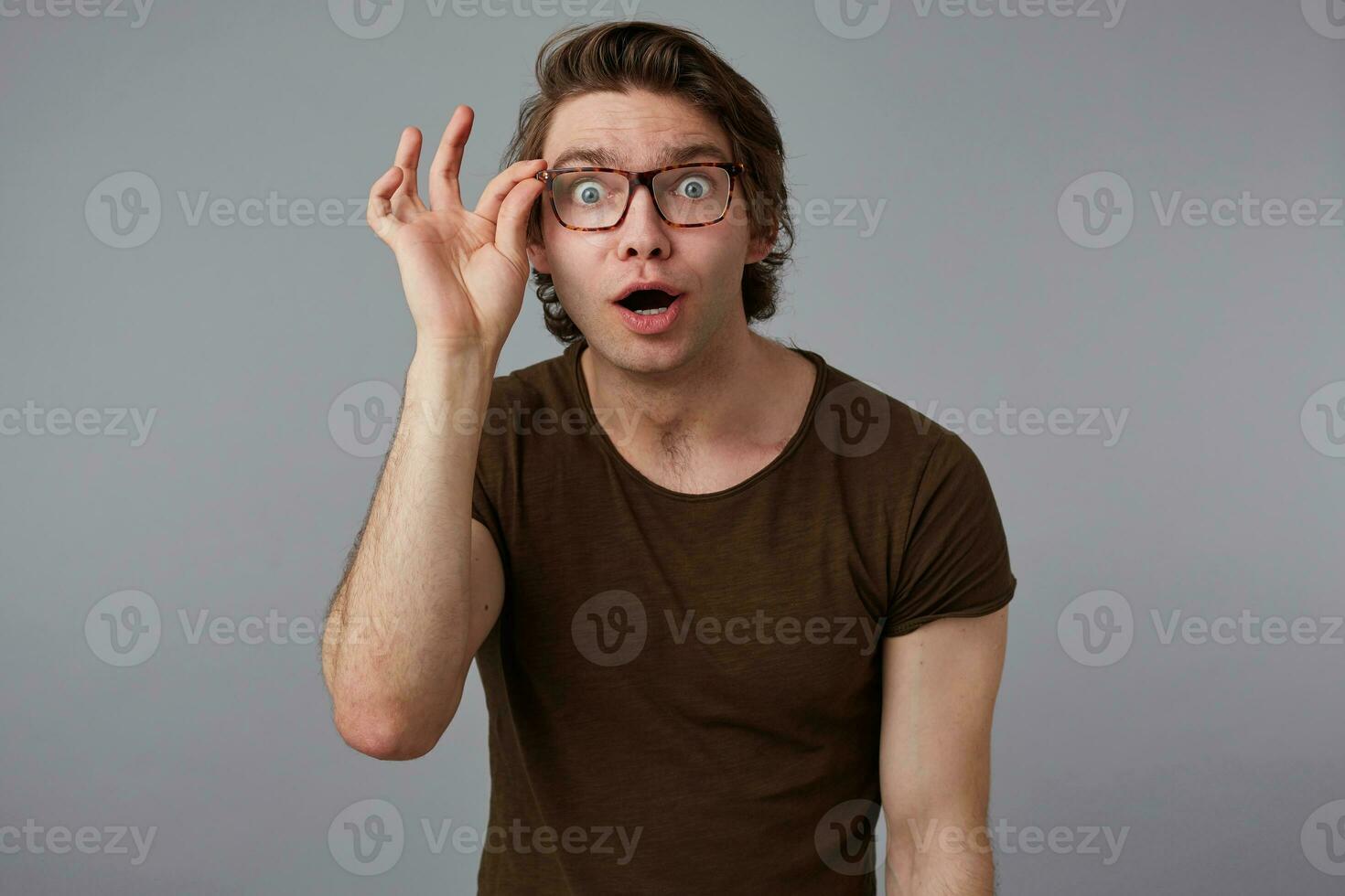 portret van jong vroeg me af Mens met bril draagt in eenvoudig t-shirt, staat over- grijs achtergrond en looks Bij de camera trog bril. foto