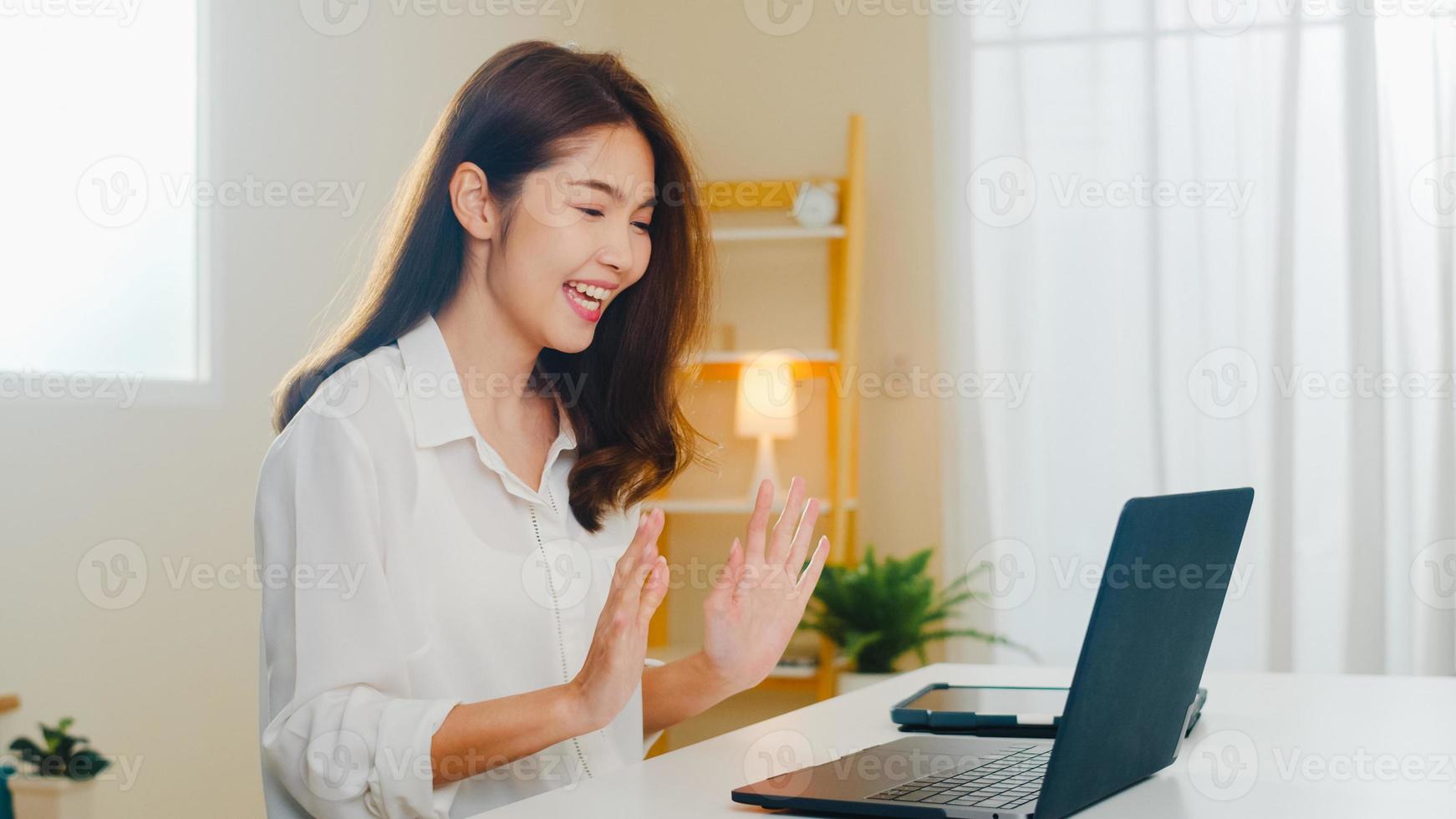 jonge Aziatische zakenvrouw die laptop videogesprek gebruikt om met een paar te praten terwijl ze vanuit huis in de woonkamer werkt. zelfisolatie, sociale afstand, quarantaine voor coronavirus in het volgende normale concept. foto