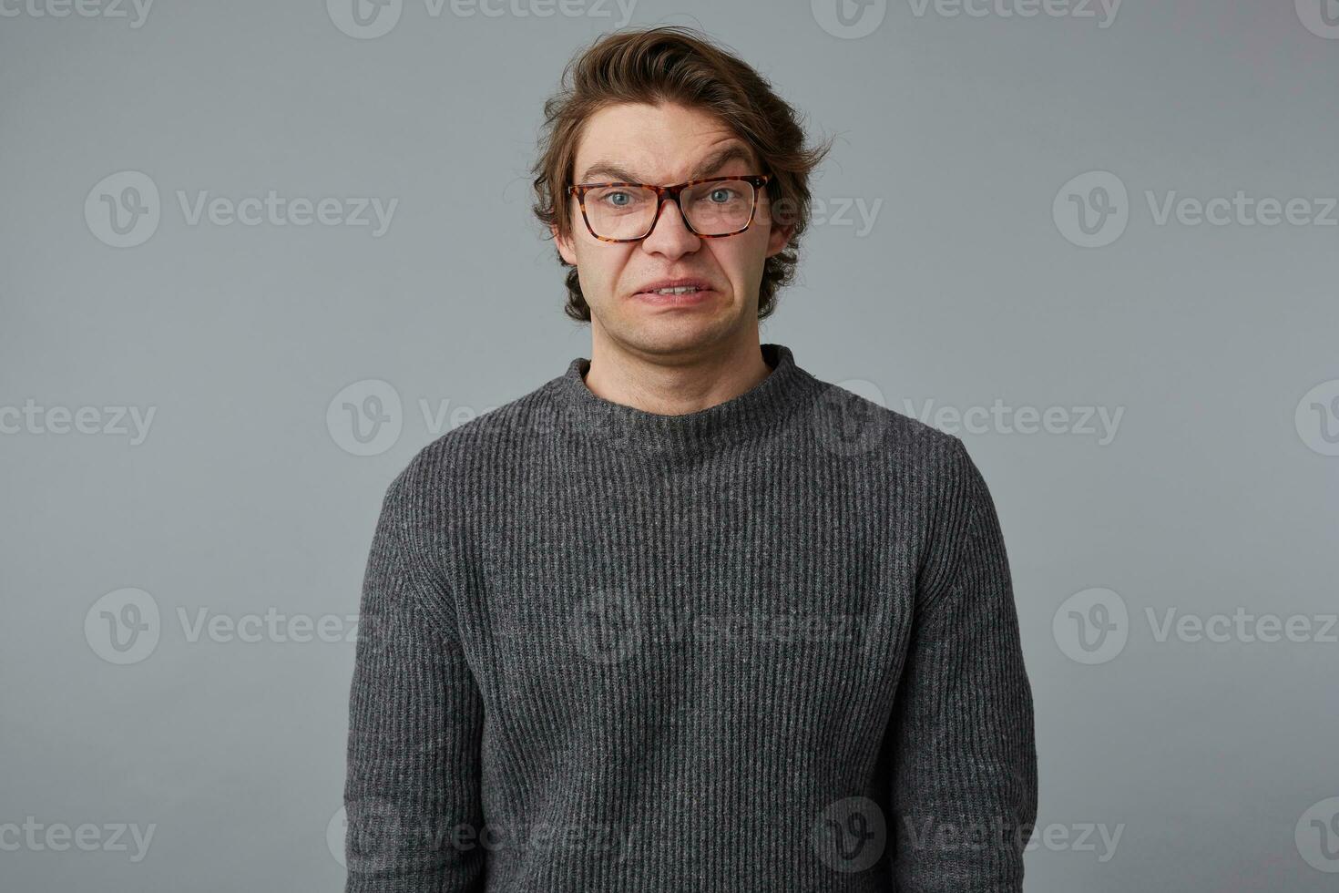 portret van jong ongelukkig Mens met bril draagt in grijs trui, staat over- grijs achtergrond en looks Bij de camera met walging. foto