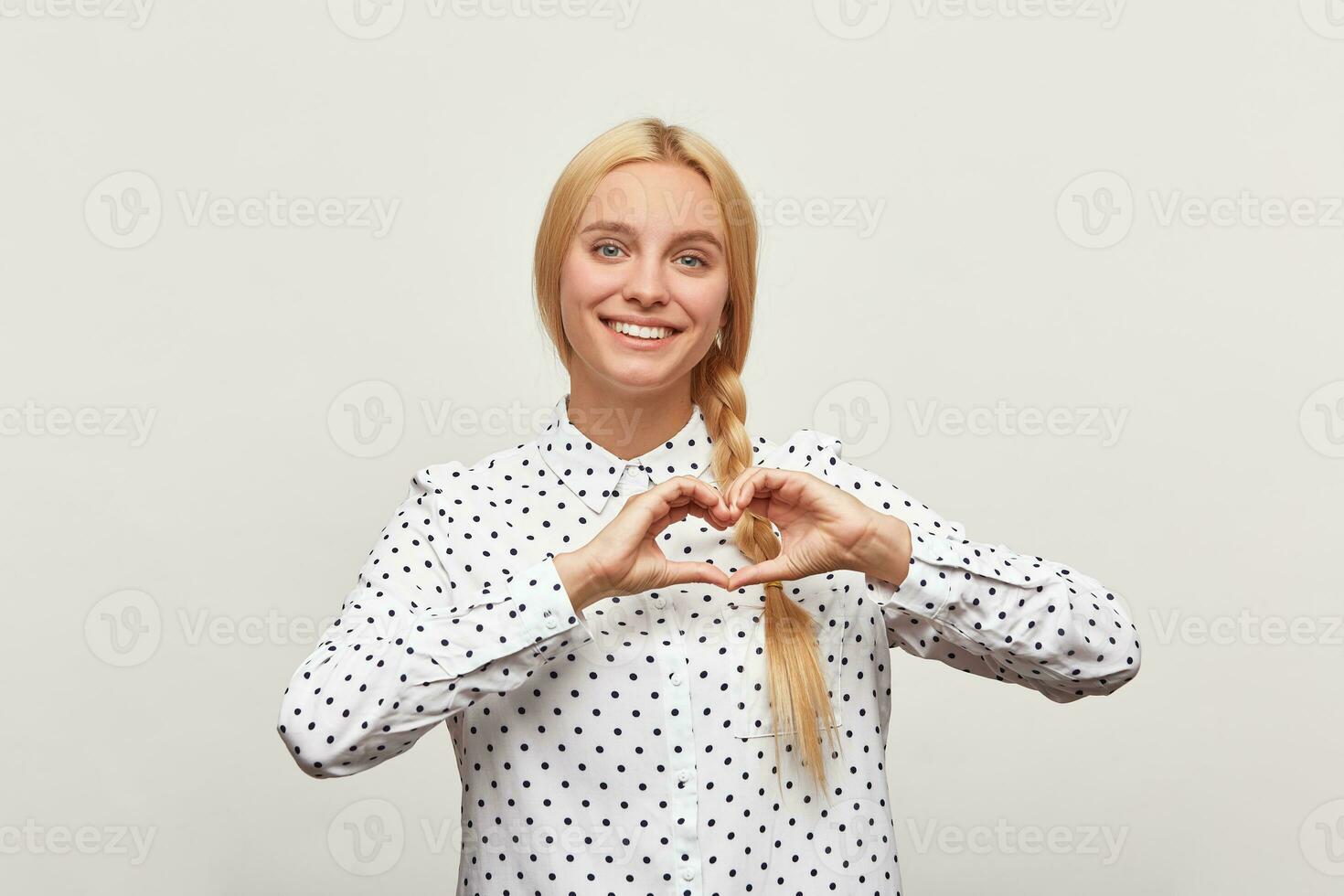 romantisch portret van inschrijving charmant vrouw Aan een wit achtergrond. meisje shows hart vorm gebaar met handen. vrouw met vlecht in overhemd tonen liefde symbool, op zoek Bij camera foto