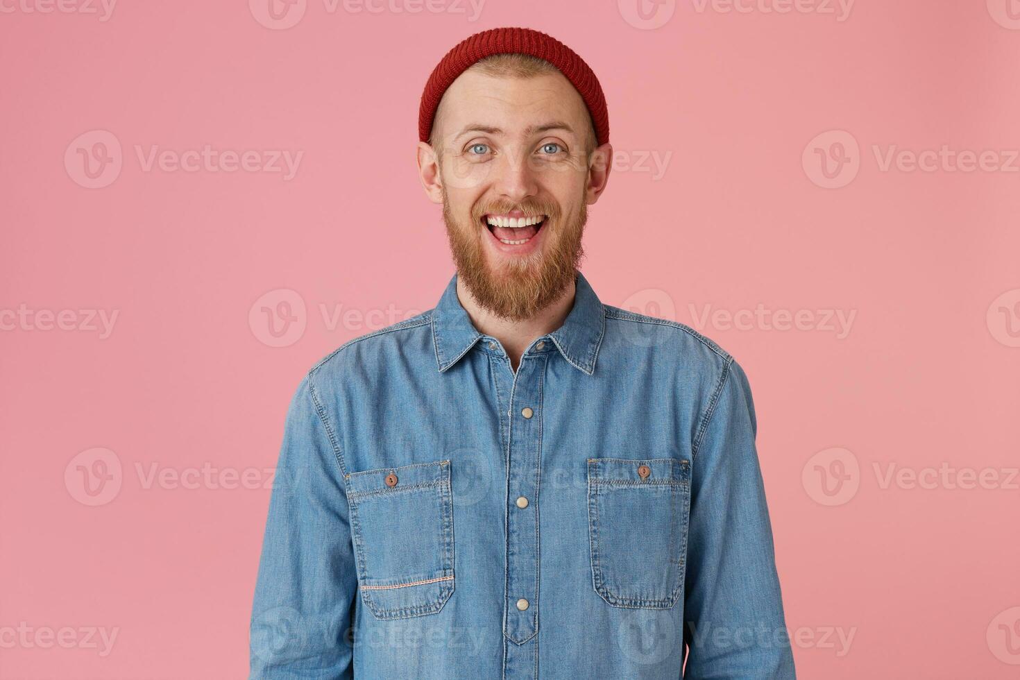 portret van lachend vent in rood hoed draagt modieus denim shirt, met rood dik baard looks gelukkig blij, heeft plezier, geïsoleerd Aan roze achtergrond foto