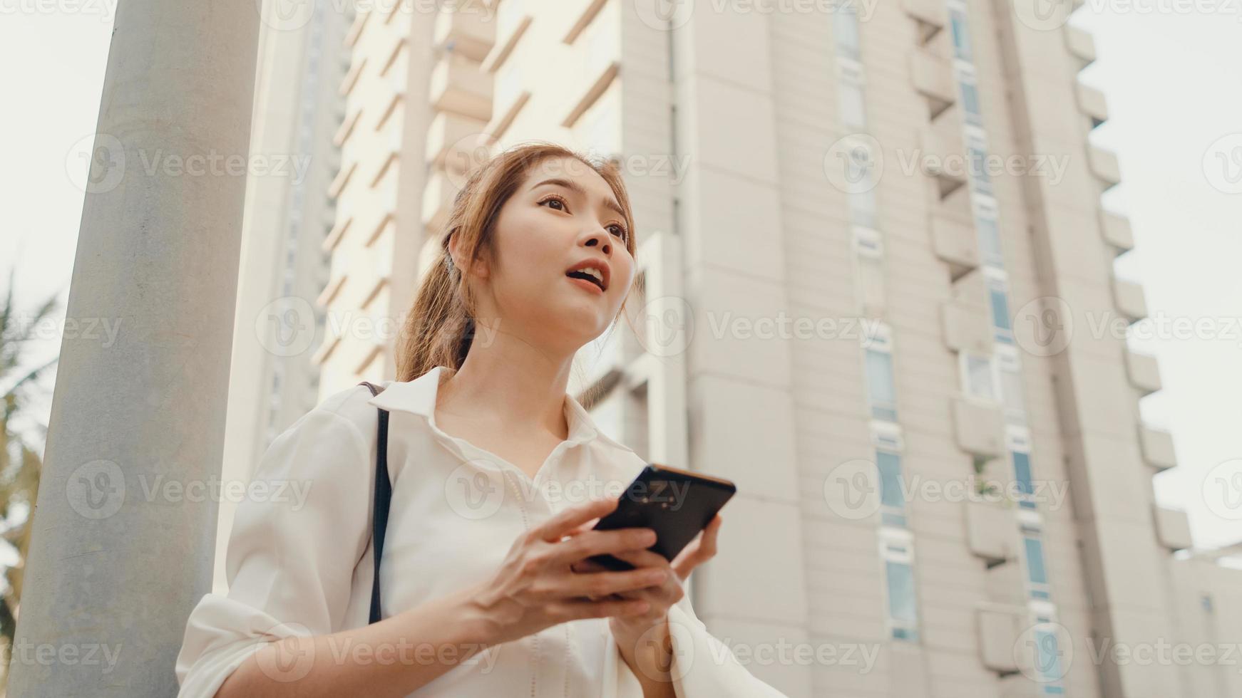succesvolle jonge azië zakenvrouw in mode kantoorkleding die op de weg een taxi neemt en een smartphone gebruikt terwijl ze buiten in de stedelijke moderne stad staat. bedrijf onderweg concept. foto