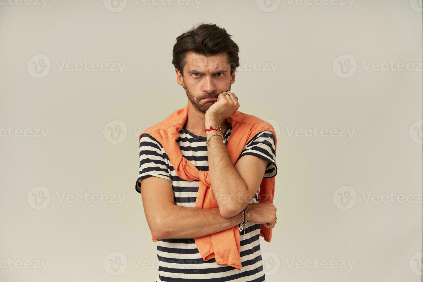 van streek Mens met brunette haar- en haren. vervelend gestreept t-shirt en oranje trui gebonden Aan schouders. slank zijn kin Aan hand. aan het kijken geconcentreerd Bij de camera geïsoleerd over- grijs achtergrond foto