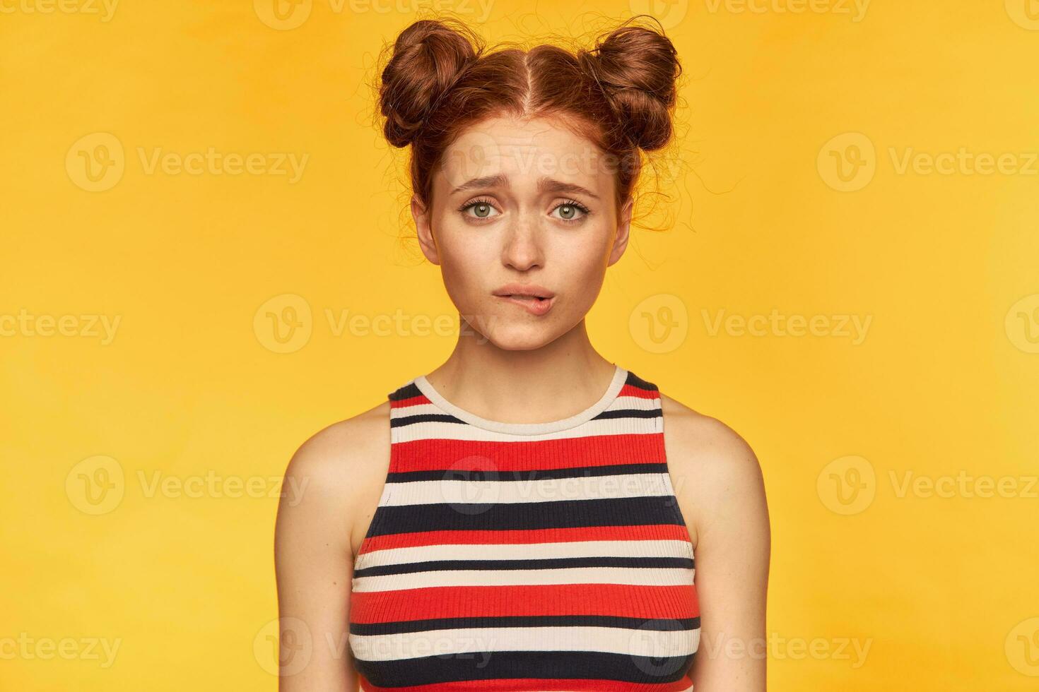 tiener- meisje, gelukkig op zoek rood haar- vrouw met twee broodjes. vervelend gestreept tank top en beet haar lip, in afwachting van wat u zullen zeggen. aan het kijken Bij de camera geïsoleerd over- geel achtergrond foto