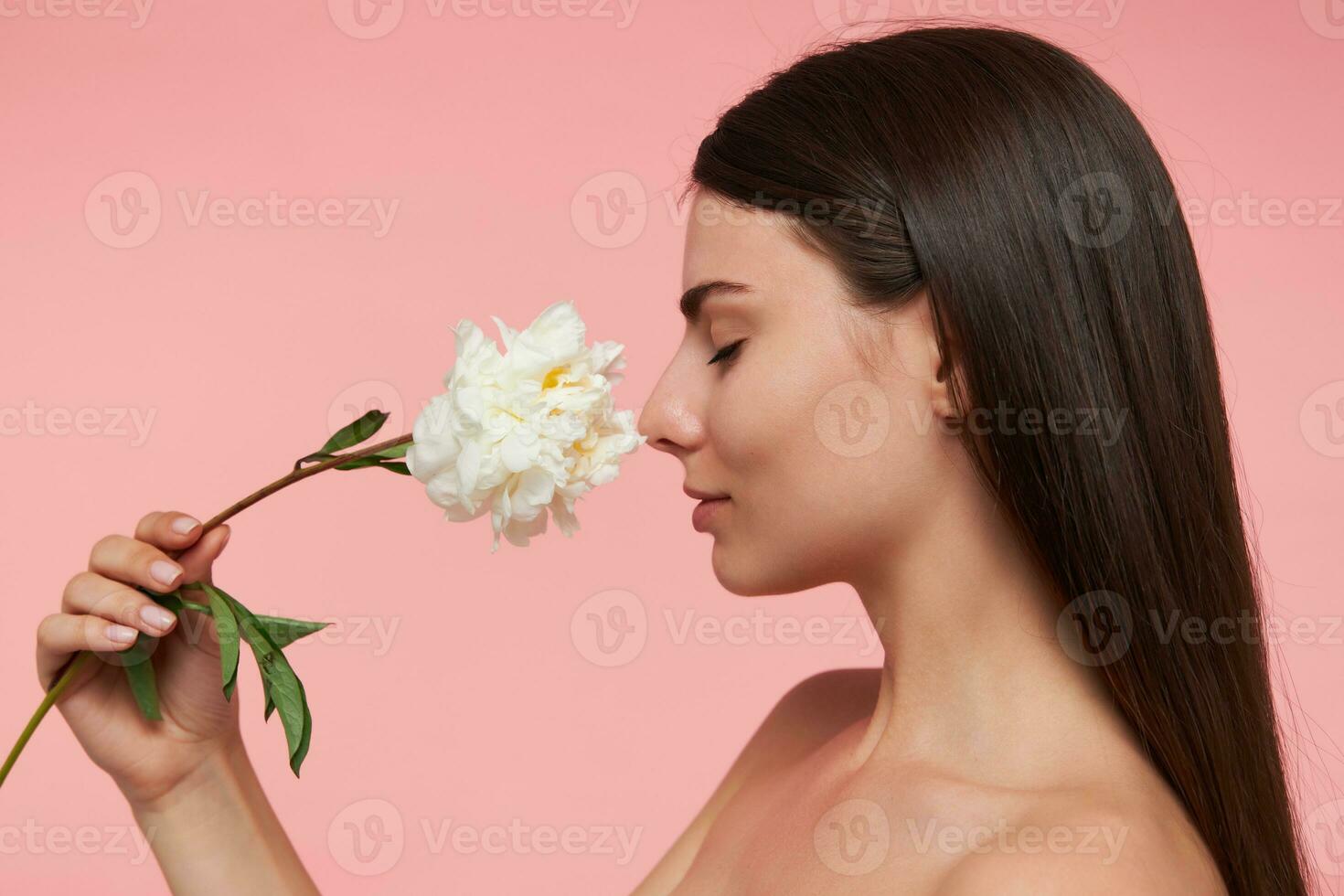 schattig op zoek vrouw met Gesloten ogen, brunette lang haar- en gezond huid, aanraken neus- met een bloem, ruikt aroma. detailopname. staan geïsoleerd over- pastel roze achtergrond foto