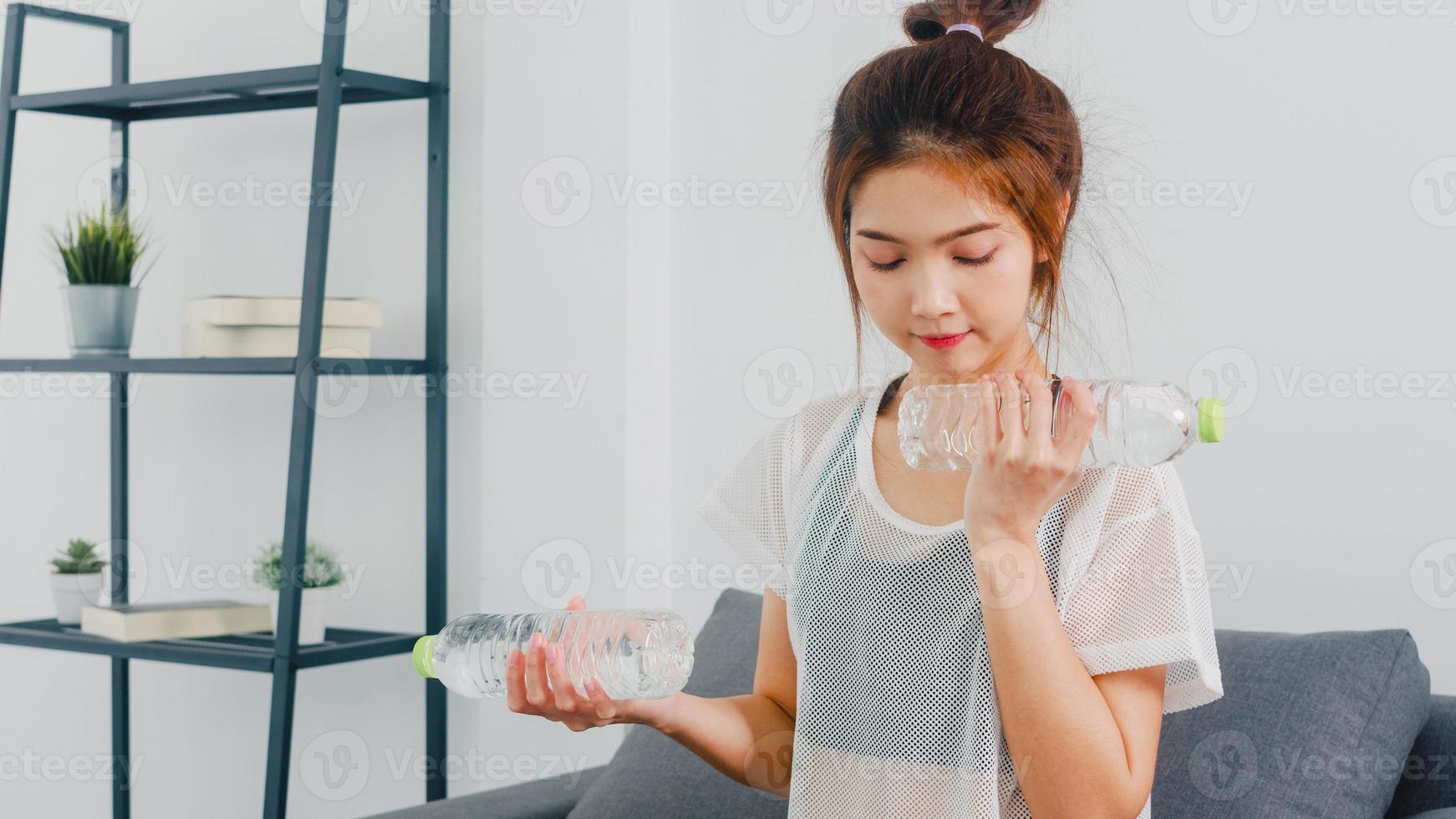 jonge Koreaanse dame in sportkleding oefeningen doen trainen met behulp van lichtgewicht halter met fles water op de bank in de woonkamer thuis. social distance, isolement tijdens het virus, blijf thuis. foto