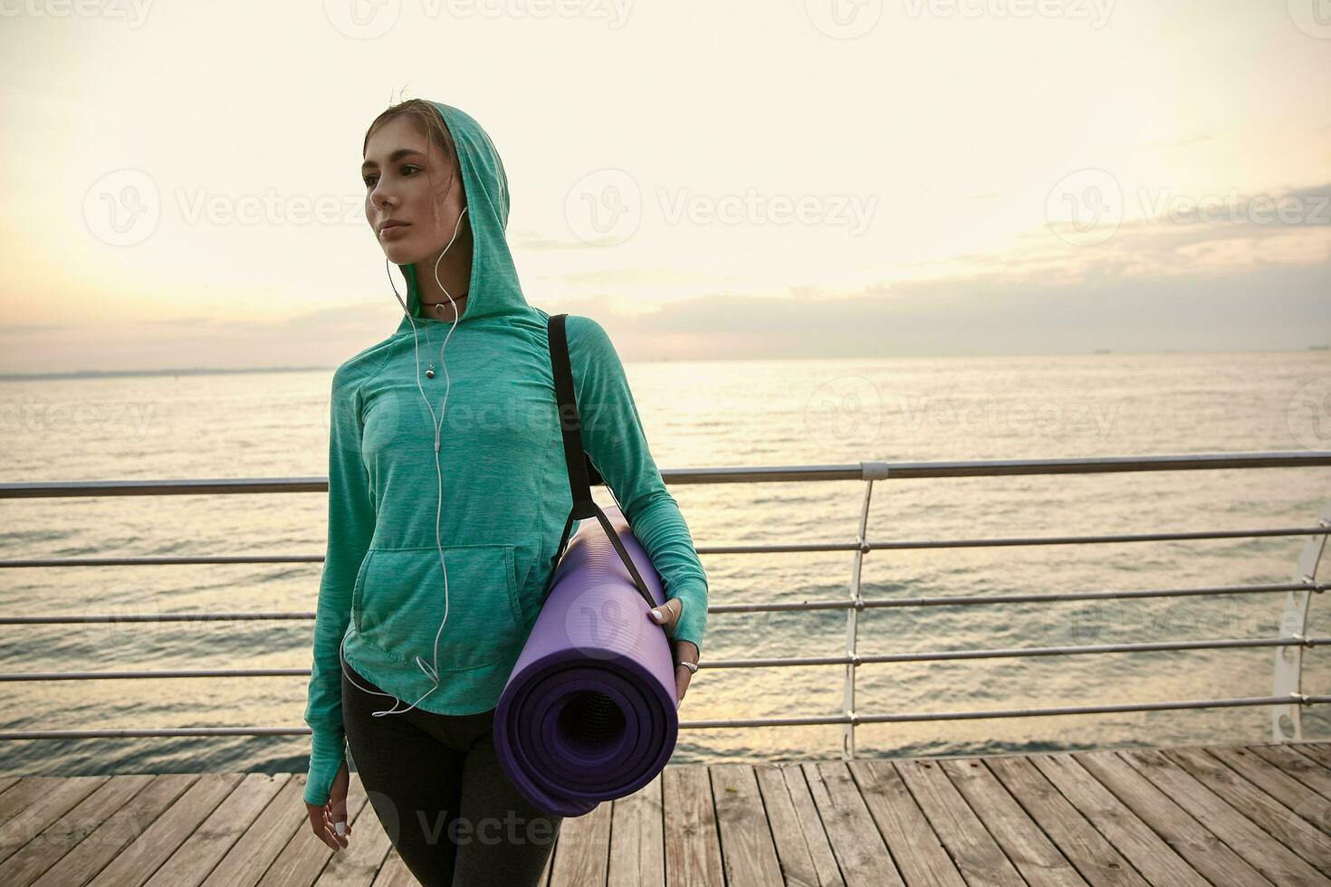jong schattig dame wandelen Bij de kust, gaan naar beoefenen yoga en maakt ochtend- uitrekken, Holding een Purper yoga mat en looks weg. Gezondheid zorg concept. foto