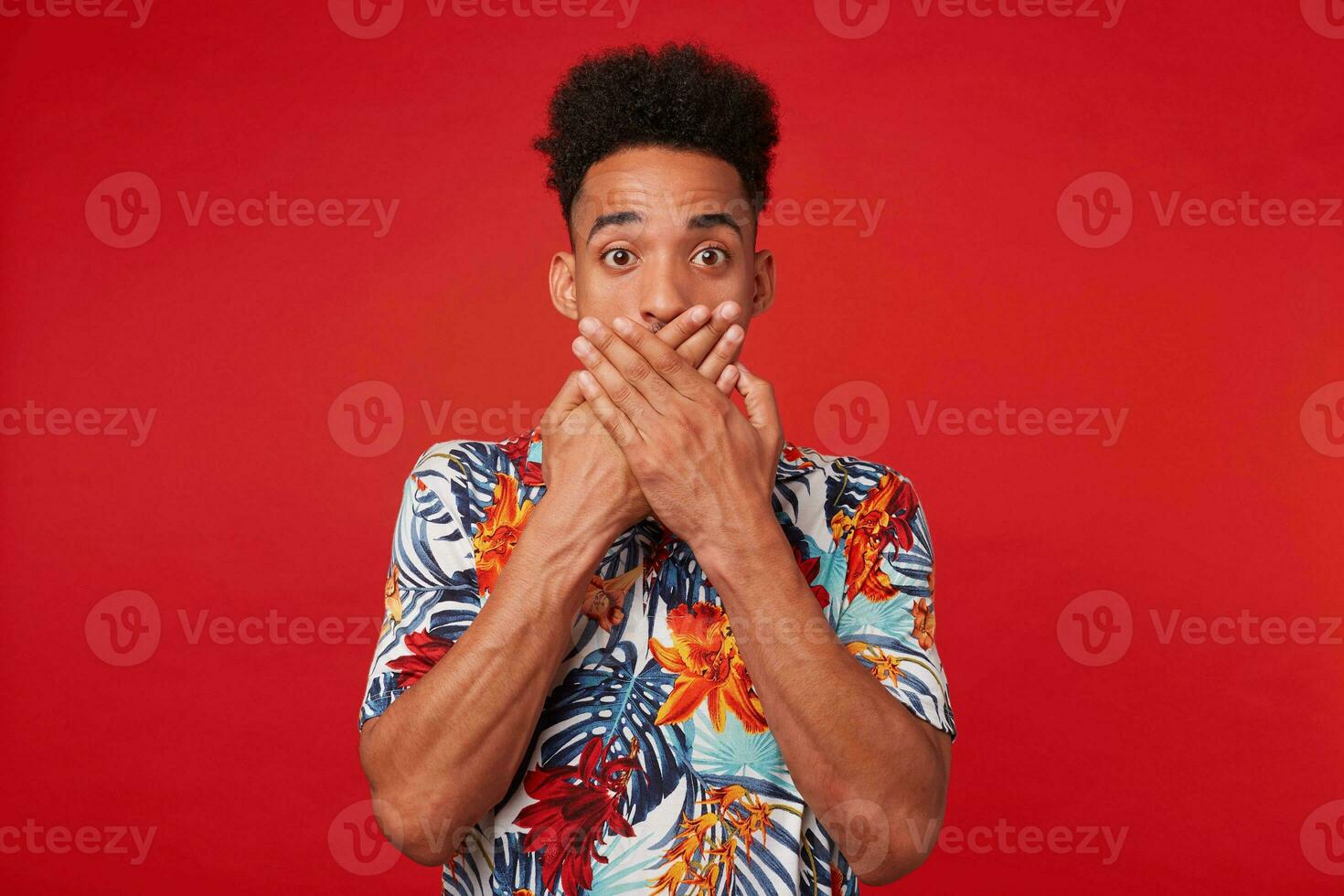 portret van jong vroeg me af Afrikaanse Amerikaans jongen, draagt in hawaiiaans shirt, looks Bij de camera met verrast uitdrukking, gedekt mond met twee handen, staat over- rood achtergrond. foto