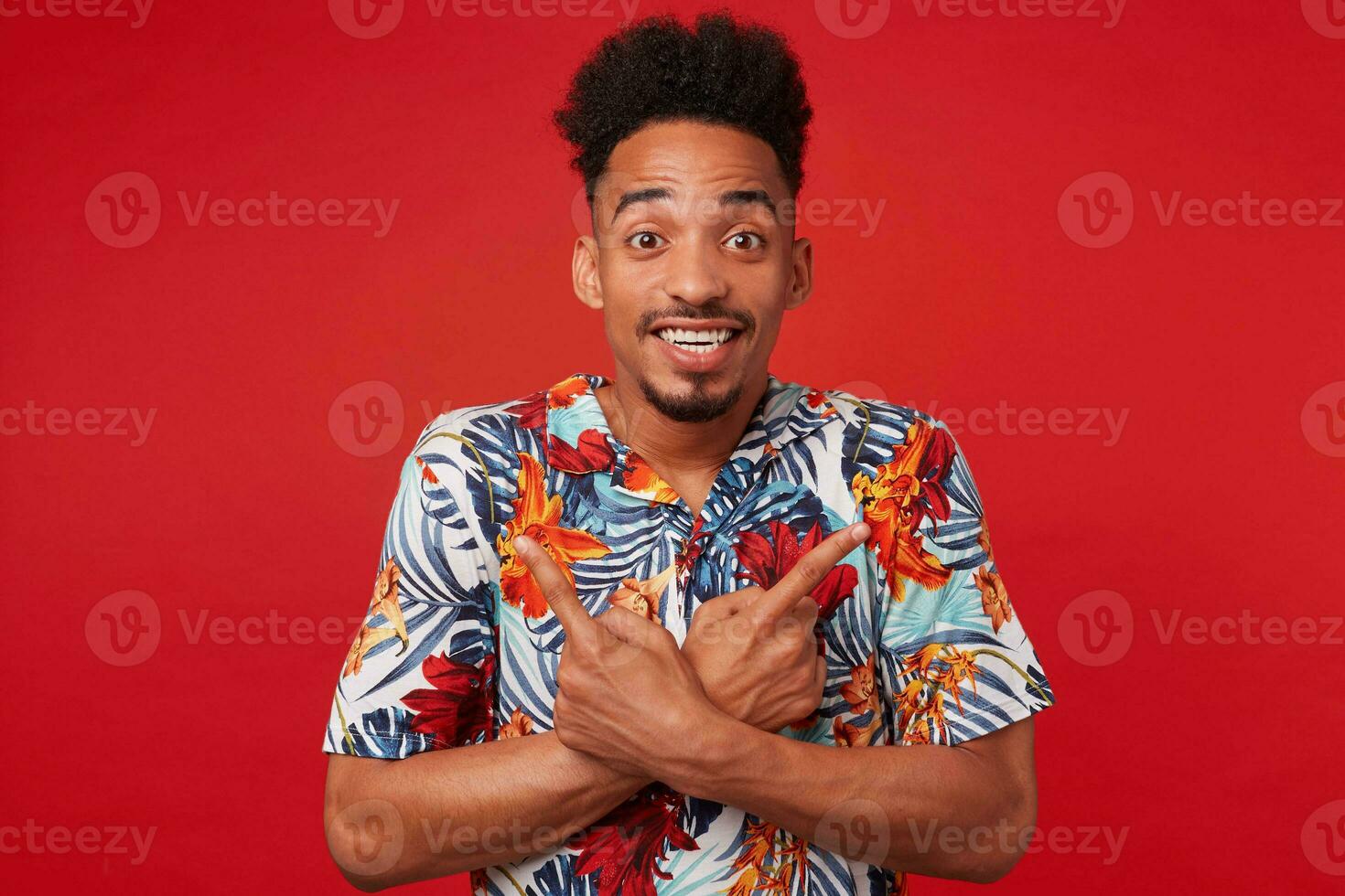 portret als jong positief Afrikaanse Amerikaans Mens in hawaiiaans shirt, looks Bij de camera met vrolijk uitdrukking, staat over- rood achtergrond en breed glimlacht, points in verschillend routebeschrijving. foto