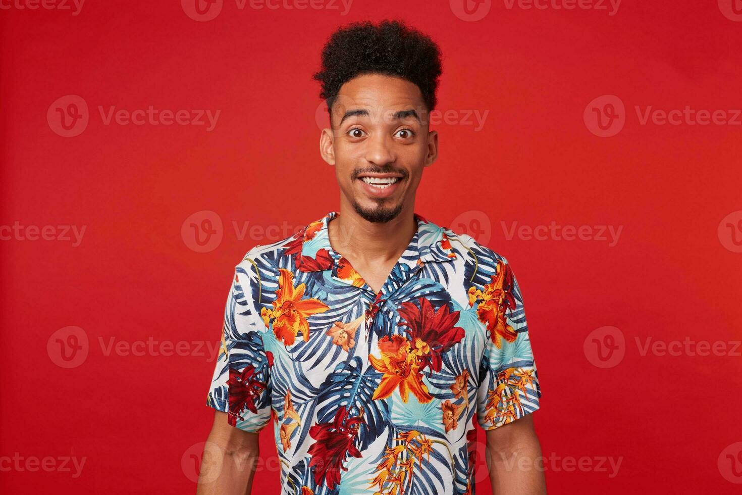 portret van gelukkig jong Afrikaanse Amerikaans jongen, draagt in hawaiiaans shirt, looks Bij de camera met vrolijk uitdrukking, staat over- rood achtergrond en breed glimlacht. foto