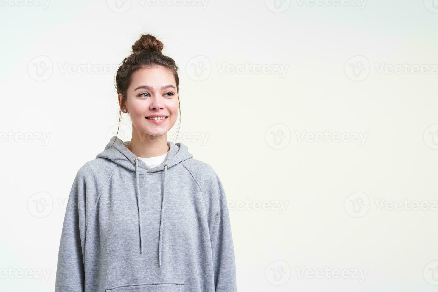 blij jong mooi vrouw vervelend haar bruin haar- in knoop terwijl poseren over- wit achtergrond in sportief kleren, op zoek positief terzijde met aangenaam glimlach foto