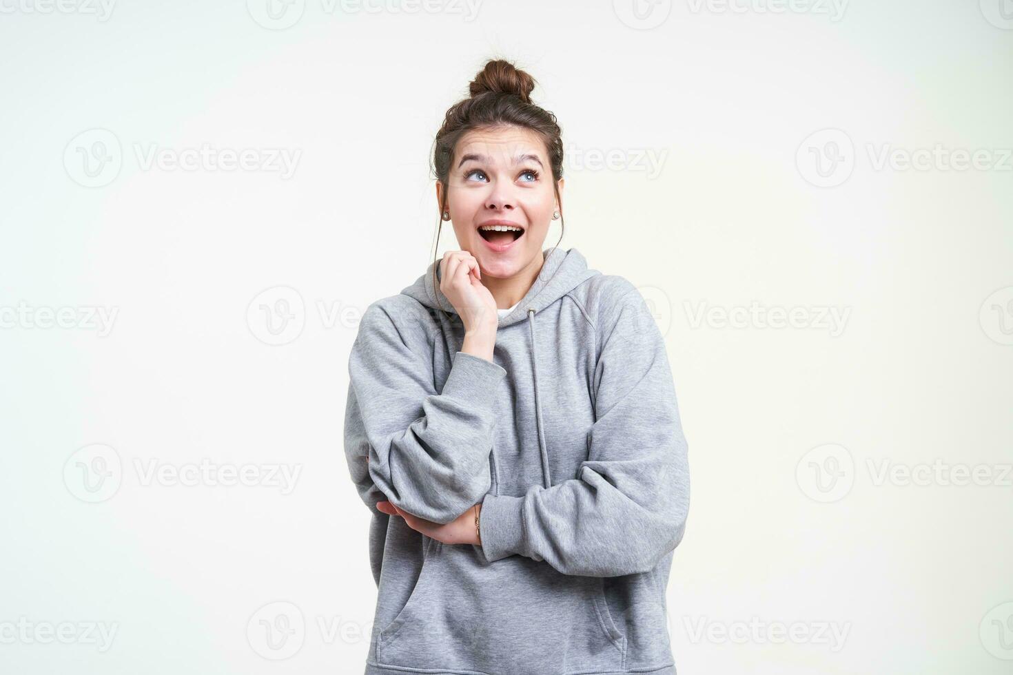 opgewonden jong mooi bruin haren vrouw met natuurlijk bedenken verhogen hand- naar haar gezicht terwijl op zoek blij omhoog met breed glimlach, geïsoleerd over- wit achtergrond foto