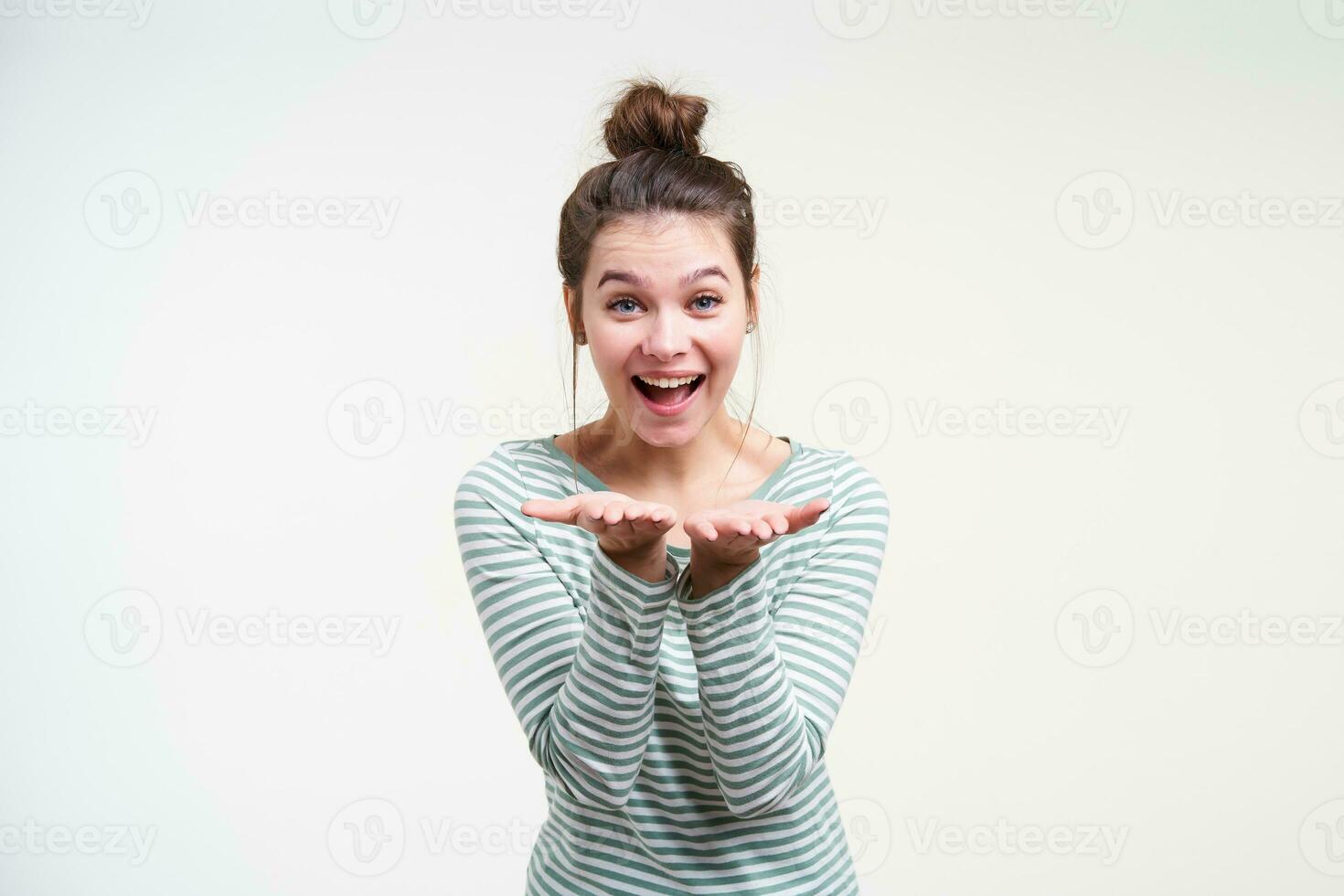 gelukkig jong mooi brunette vrouw met bun kapsel houden verheven palmen in voorkant van haarzelf en op zoek blij Bij camera, staand over- wit achtergrond foto